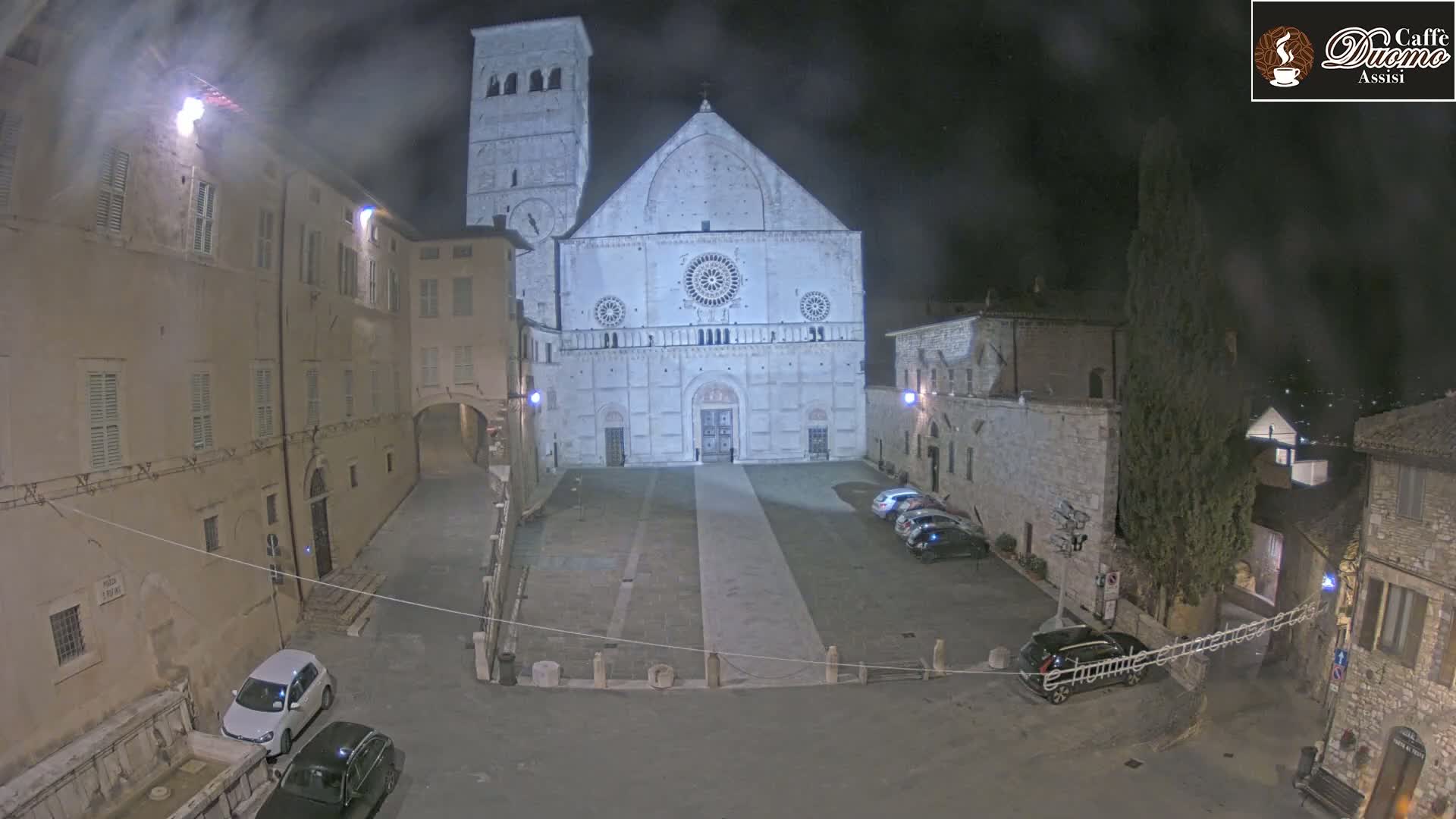 A large, light-colored cathedral is visible at night in a town square, with several cars parked around the perimeter under a hazy sky.