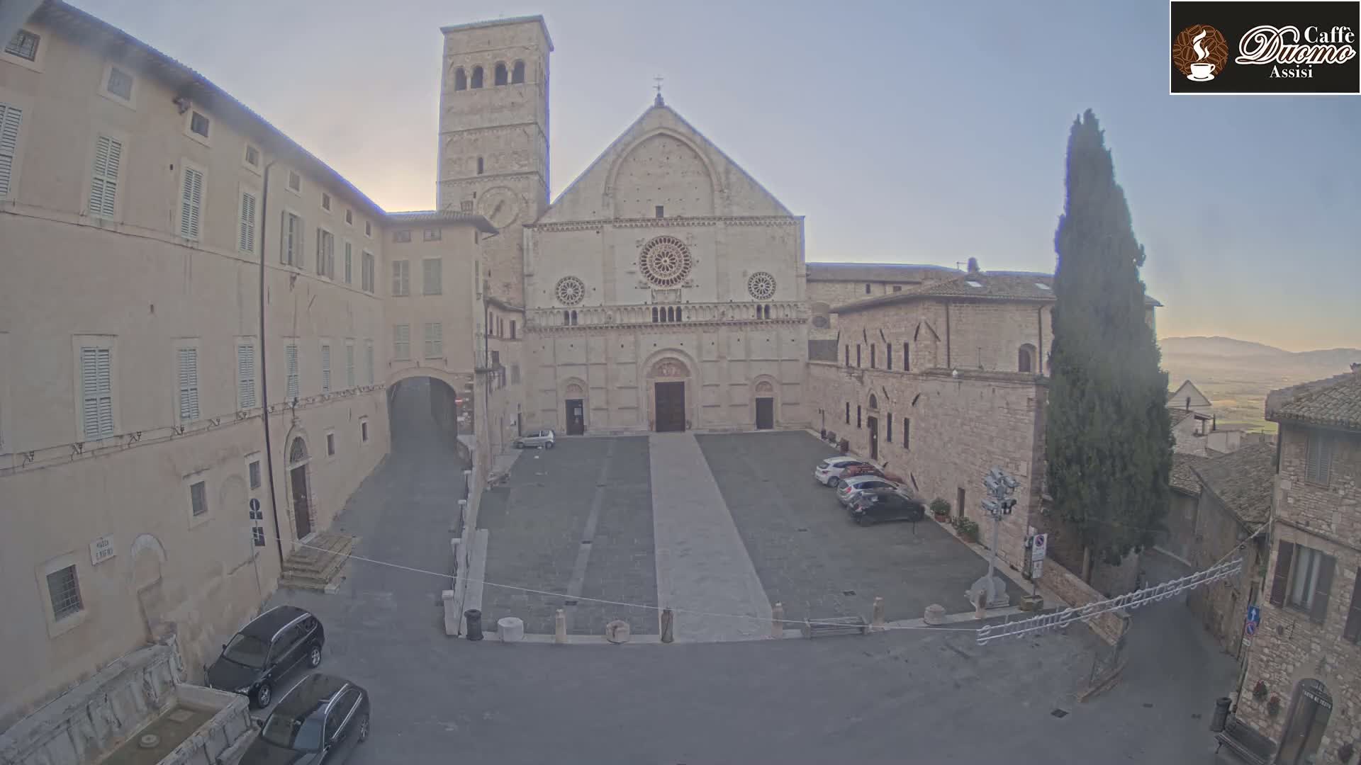 A large stone cathedral stands in a town square under a hazy, light-colored sky, with several cars parked nearby and buildings surrounding the square.