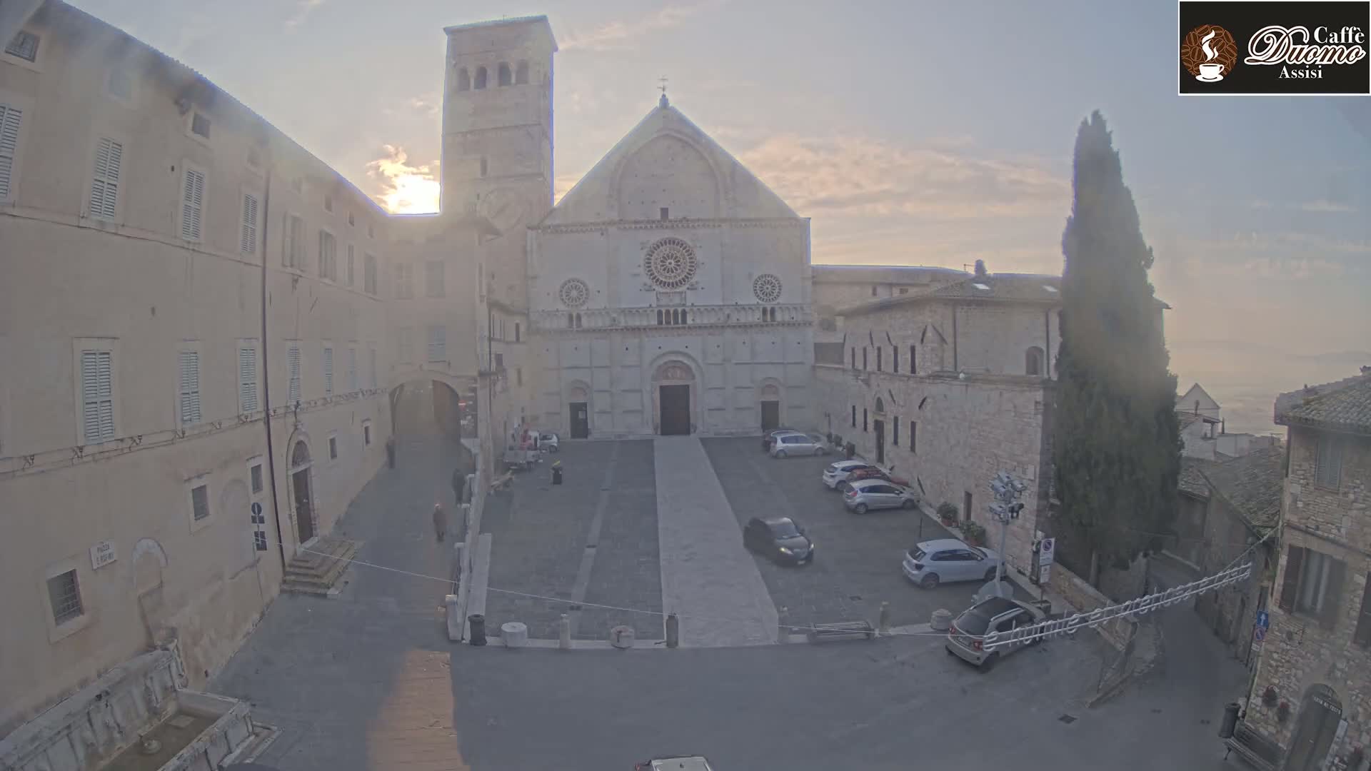 A large stone cathedral sits in a town square with several cars parked, under a hazy, sunrise sky.