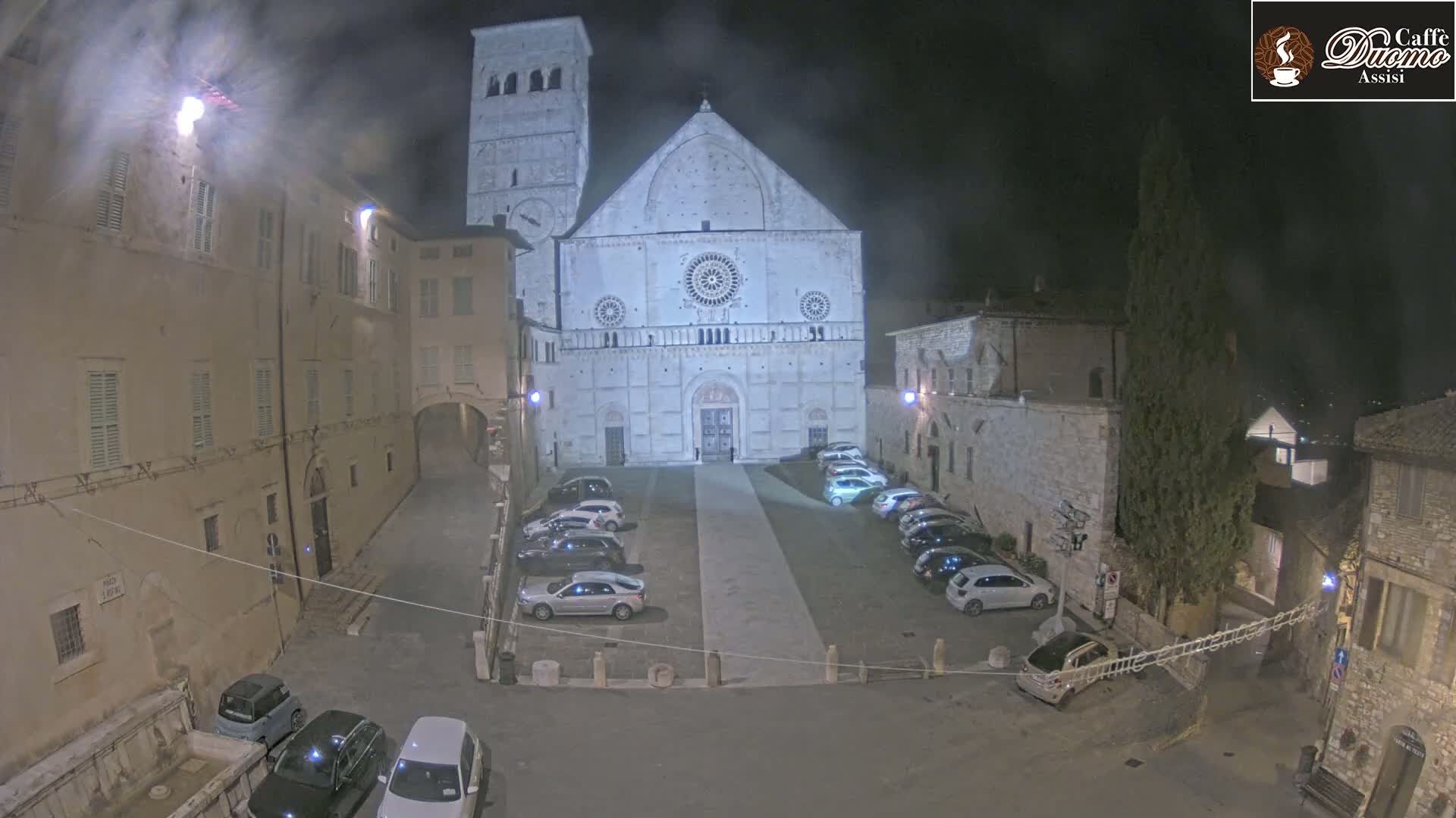 A nighttime view of a town square with cars parked in front of a large stone cathedral under hazy or foggy conditions.