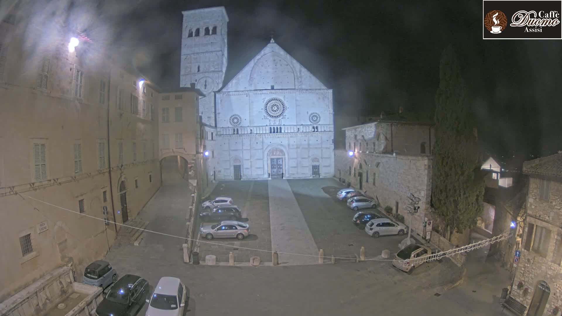 A large, light-colored cathedral stands in a dimly lit, empty town square at night, surrounded by older stone buildings and a few parked cars.