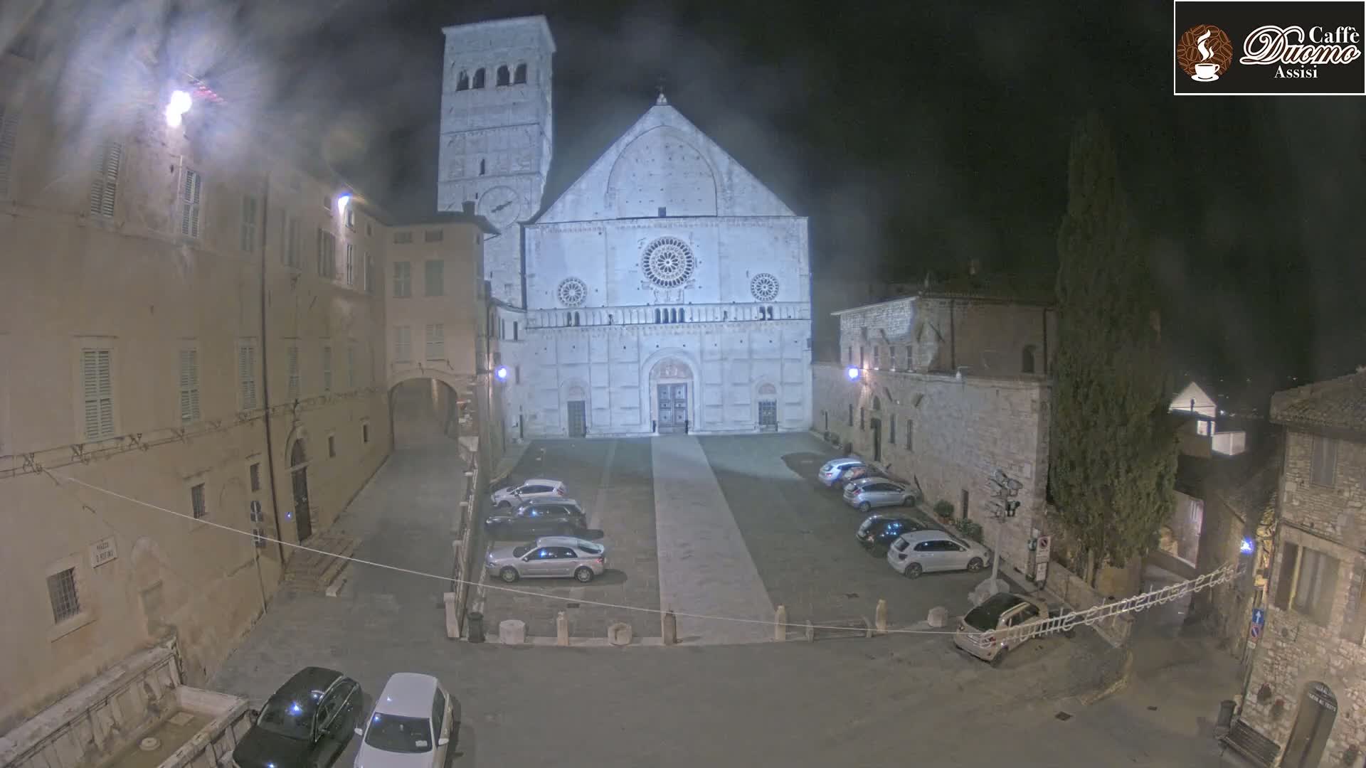 A large, light-colored cathedral is visible at night in a town square with several cars parked, under hazy or foggy conditions.