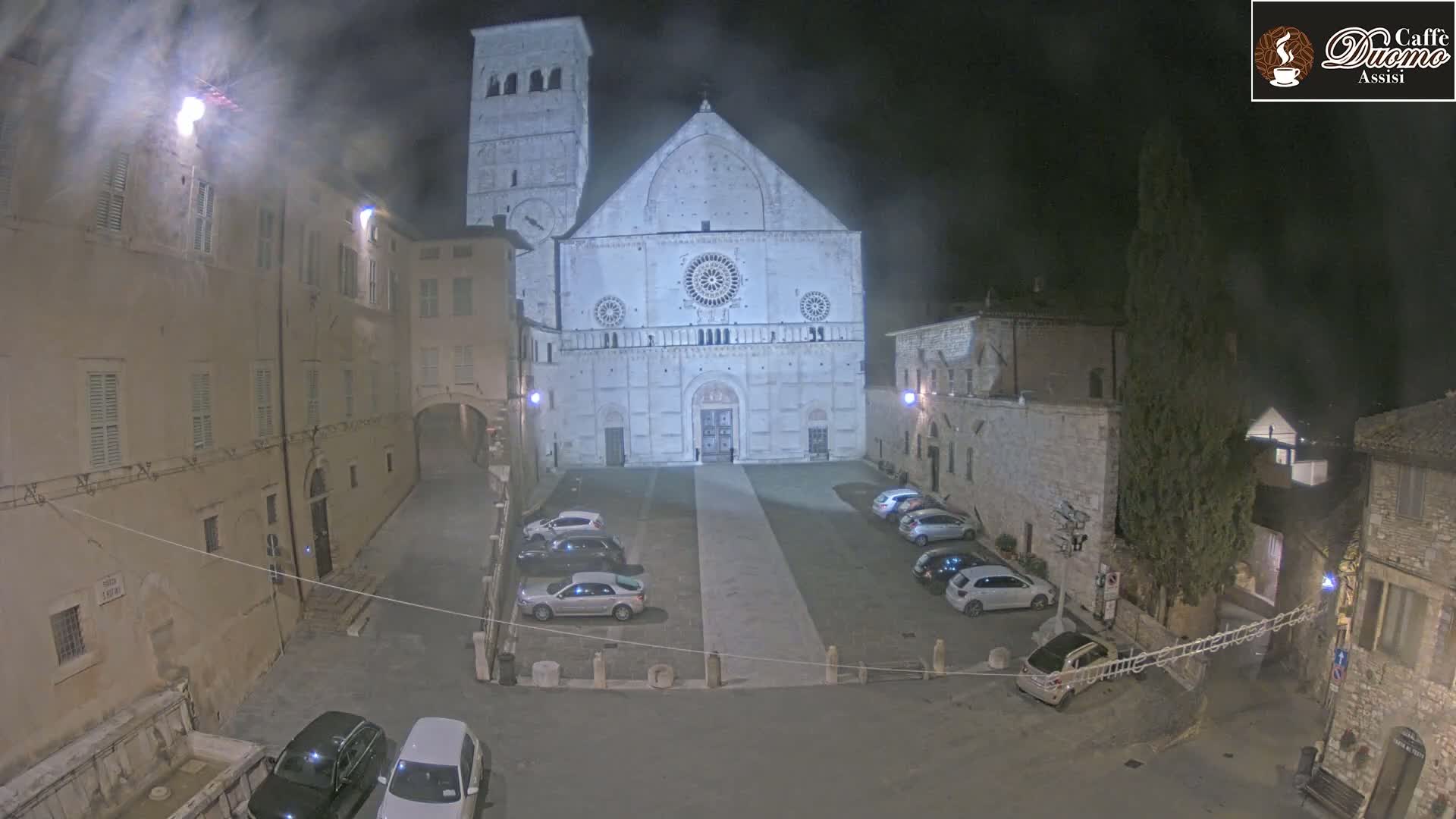 A large, light-colored cathedral stands in a dimly lit, empty square at night, surrounded by stone buildings and a few parked cars.