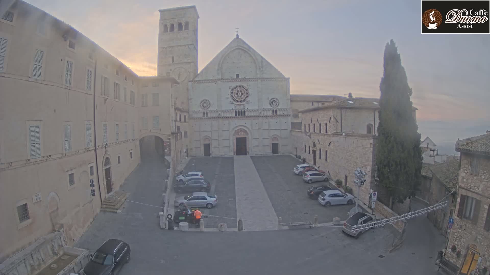 A large stone cathedral sits in a plaza with several cars parked, overlooked by adjacent buildings under a hazy, overcast sky.