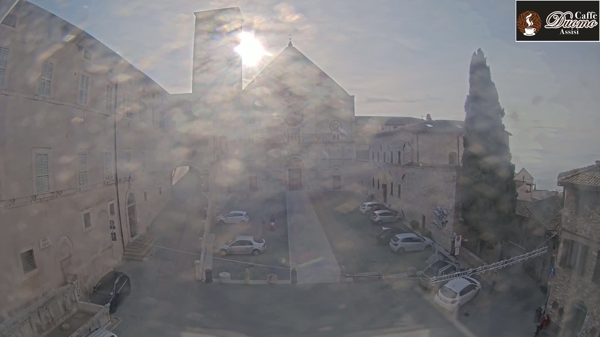 A hazy sun shines on a plaza with parked cars, surrounded by aged stone buildings, including a large cathedral.