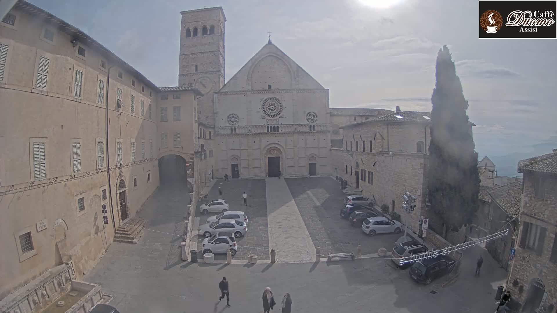 A large stone church sits in a plaza with several cars parked and a few pedestrians walking, under a hazy, sunny sky.
