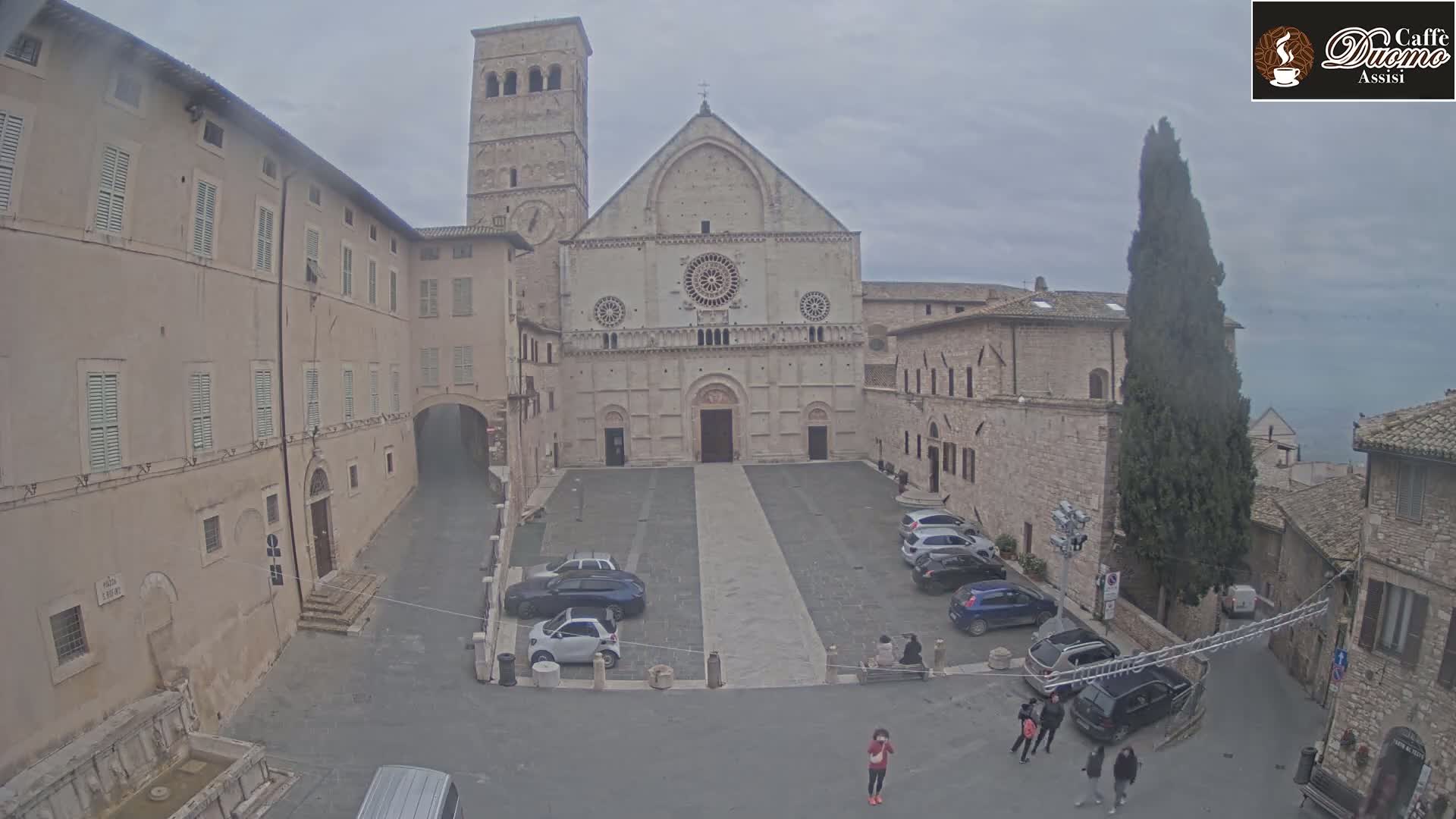 A large stone cathedral stands in a town square, with several cars parked and a few people walking around on an overcast day.