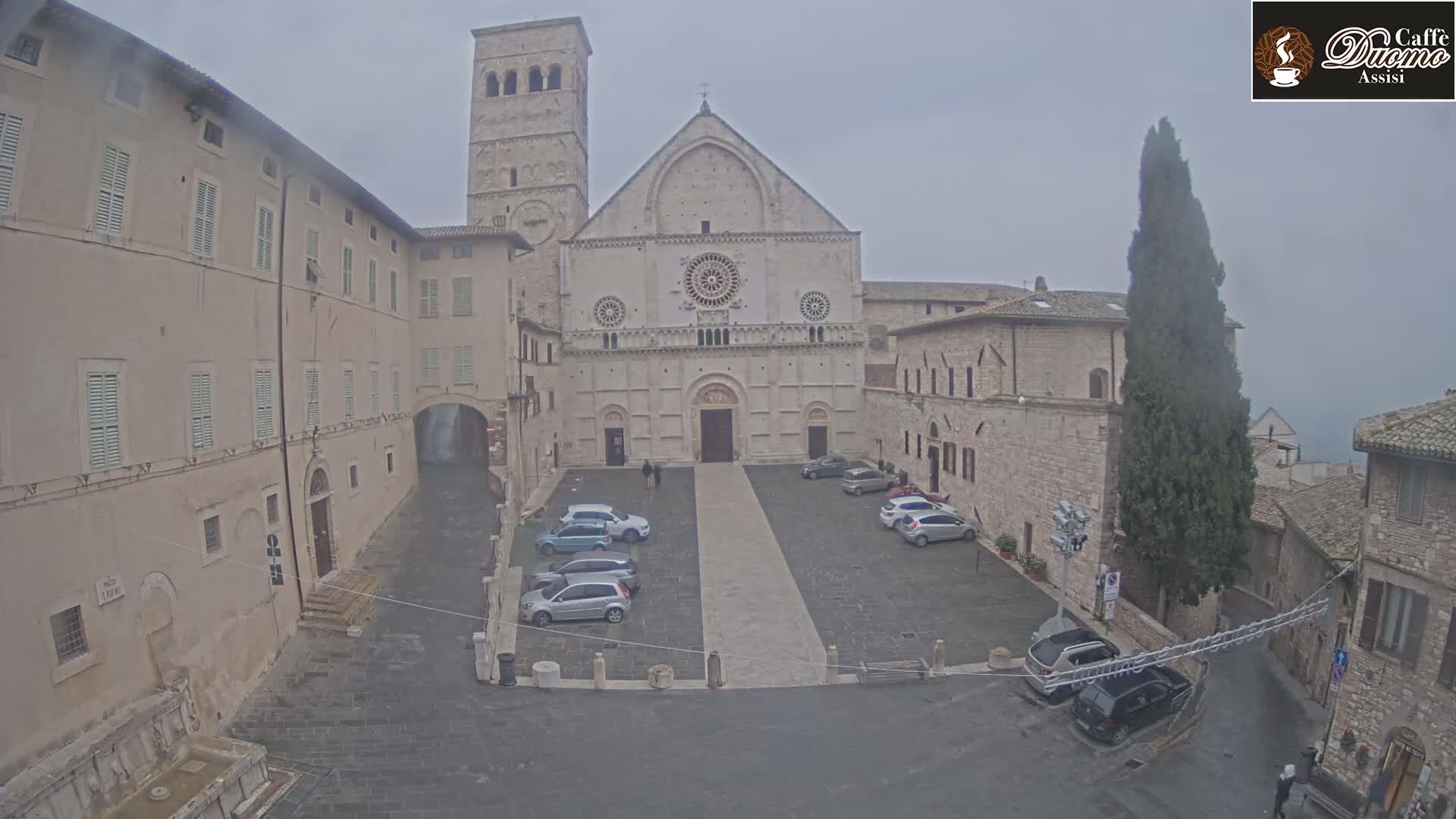A stone cathedral and surrounding buildings are visible in a square with several cars parked, on an overcast day.