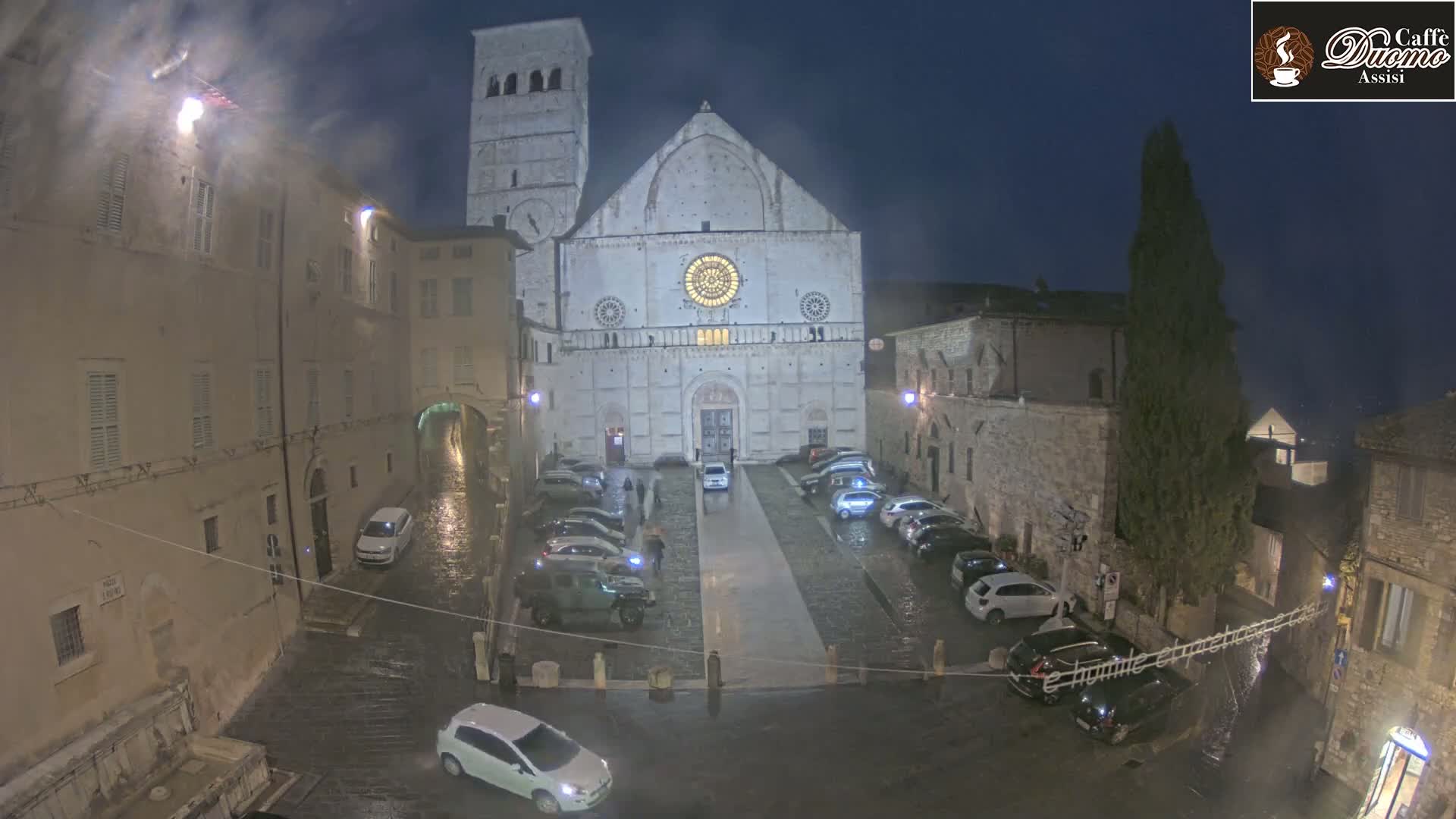 A large stone cathedral is surrounded by a wet, cobblestone square with parked cars under a dark, nighttime sky.