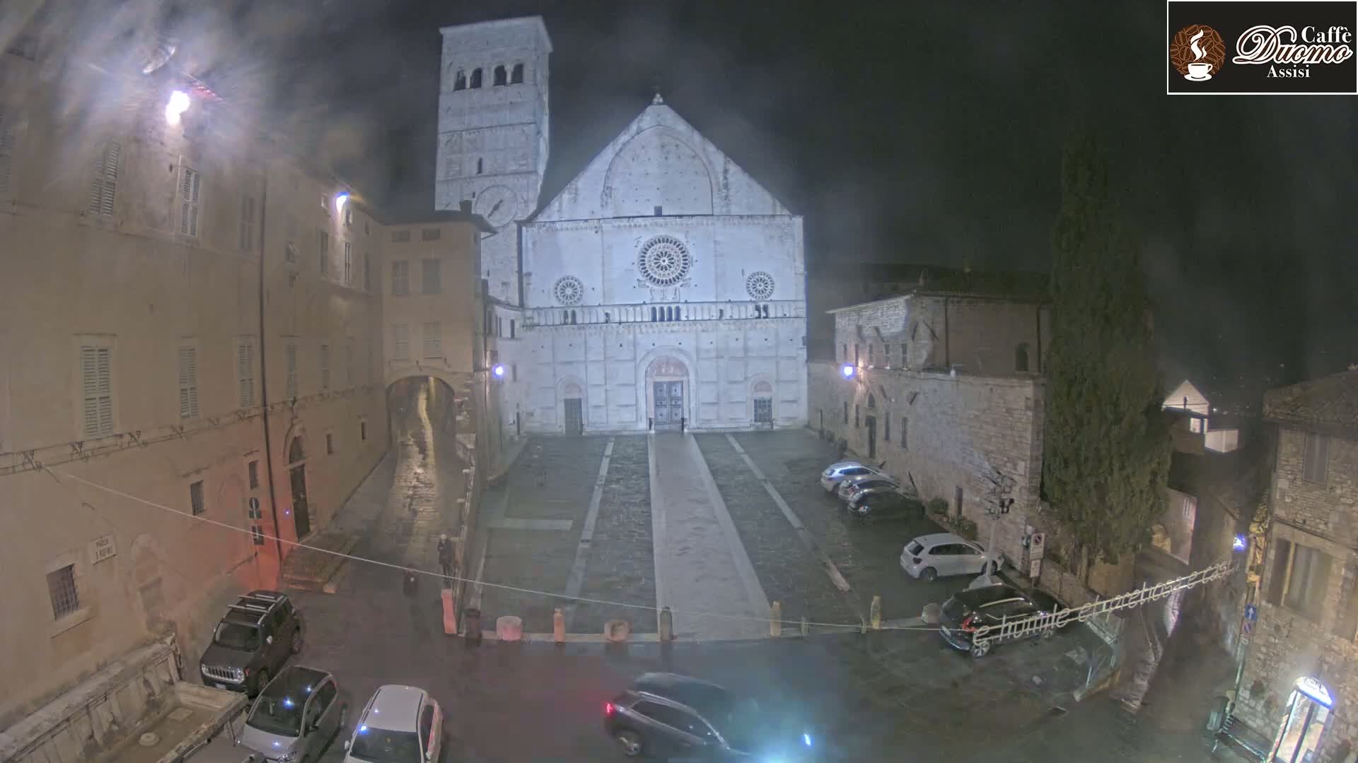 A large, light-colored cathedral stands in a town square at night, under a misty sky, surrounded by parked cars and older buildings.