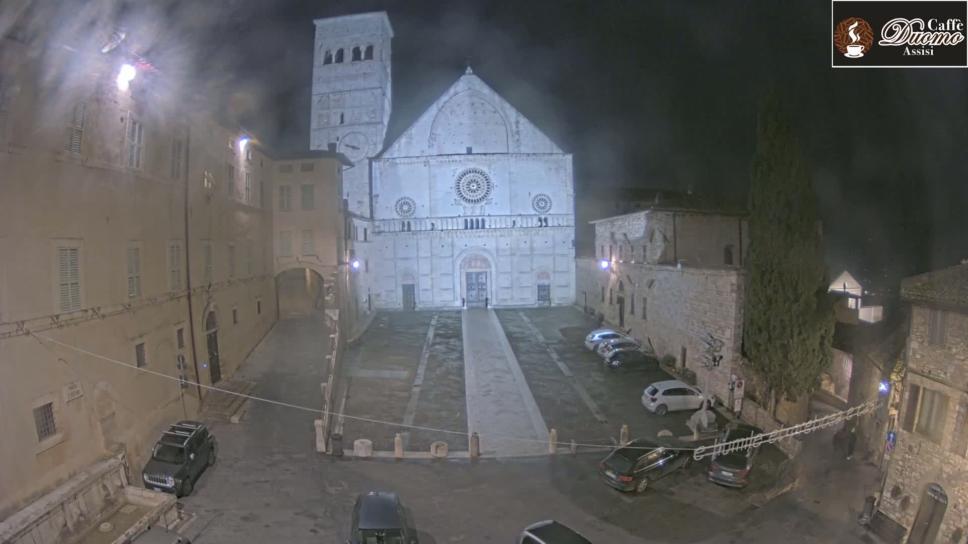 A large stone cathedral is illuminated at night in a town square with several cars parked, under a cloudy, dark sky.