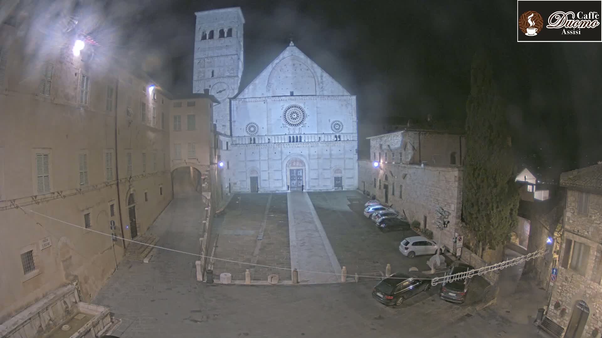 A large, light-colored cathedral is the focal point of a nighttime square, surrounded by stone buildings and a few parked cars in hazy conditions.
