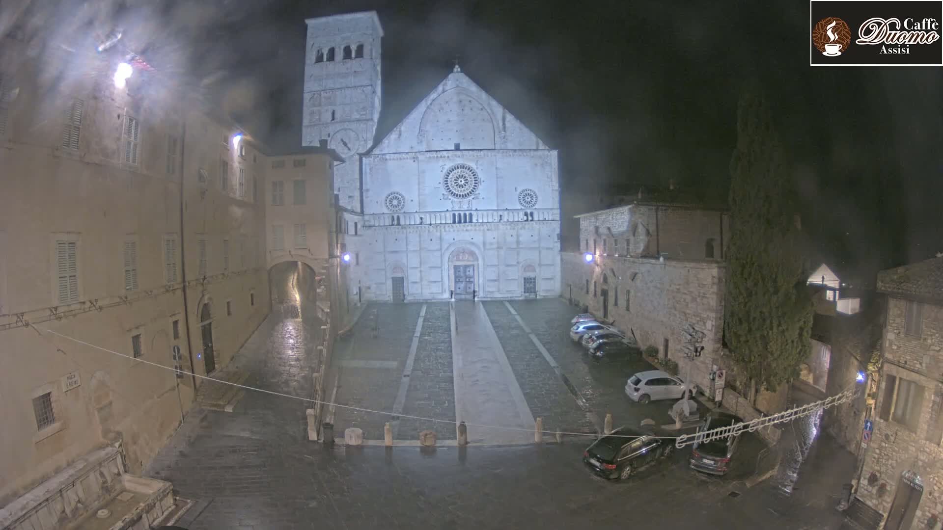 A large cathedral stands in a town square at night during a light rain, with cars parked along the sides and buildings lining the square.