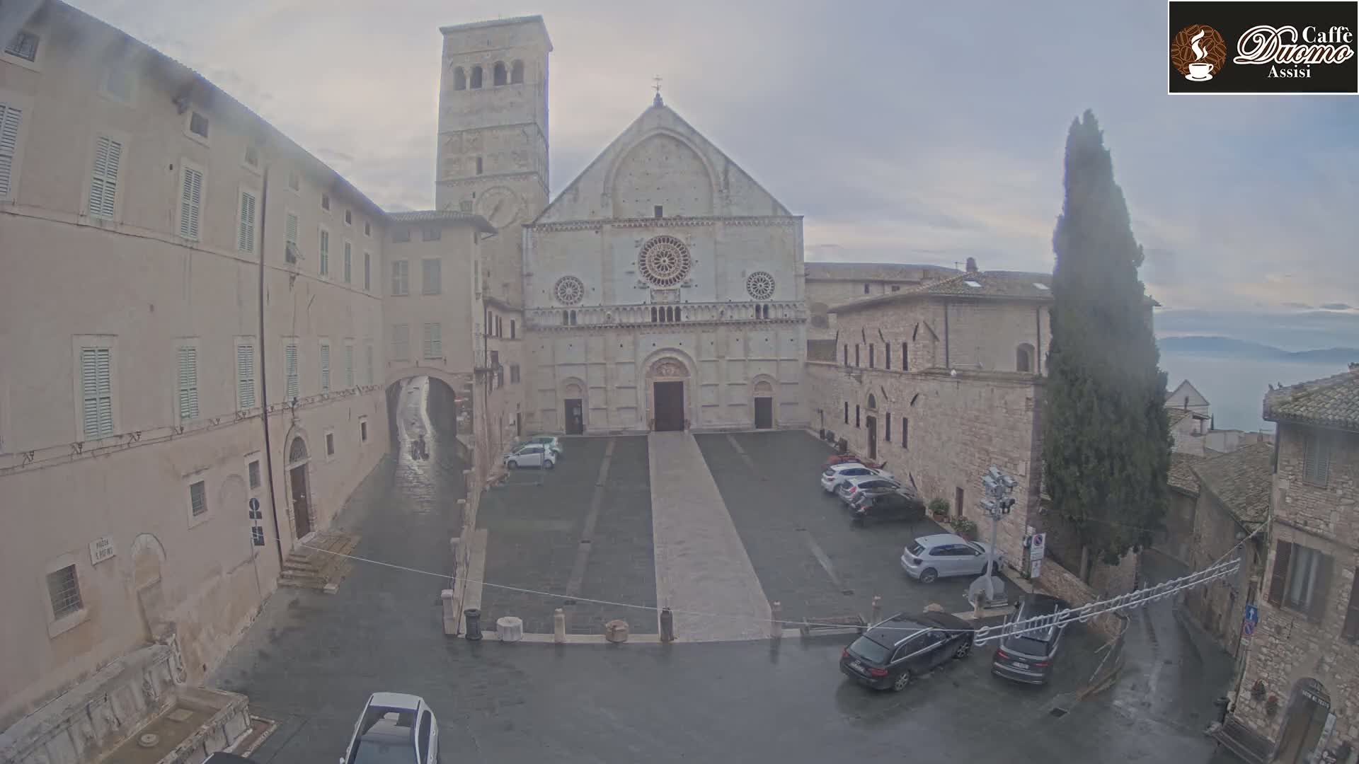 A large stone cathedral is situated in a town square with several parked cars, under an overcast sky with light rain.