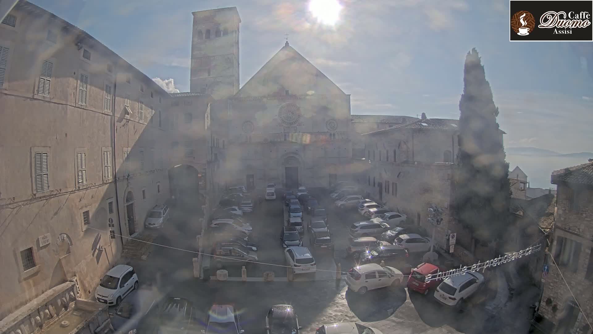 A sunny day reveals a courtyard filled with parked cars, surrounded by ancient stone buildings, including a large church with a rose window.