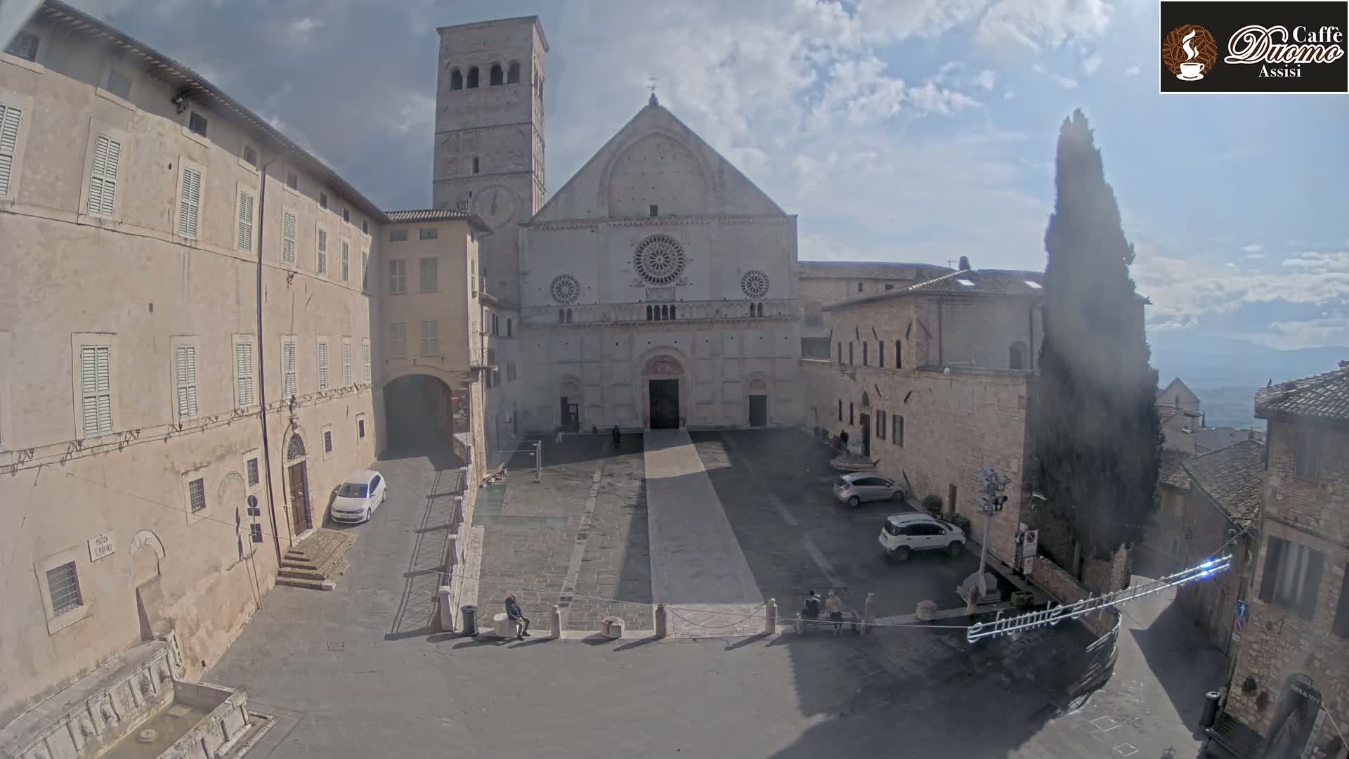 A large stone cathedral stands in a town square on a partly cloudy day, surrounded by other stone buildings and a few parked cars.