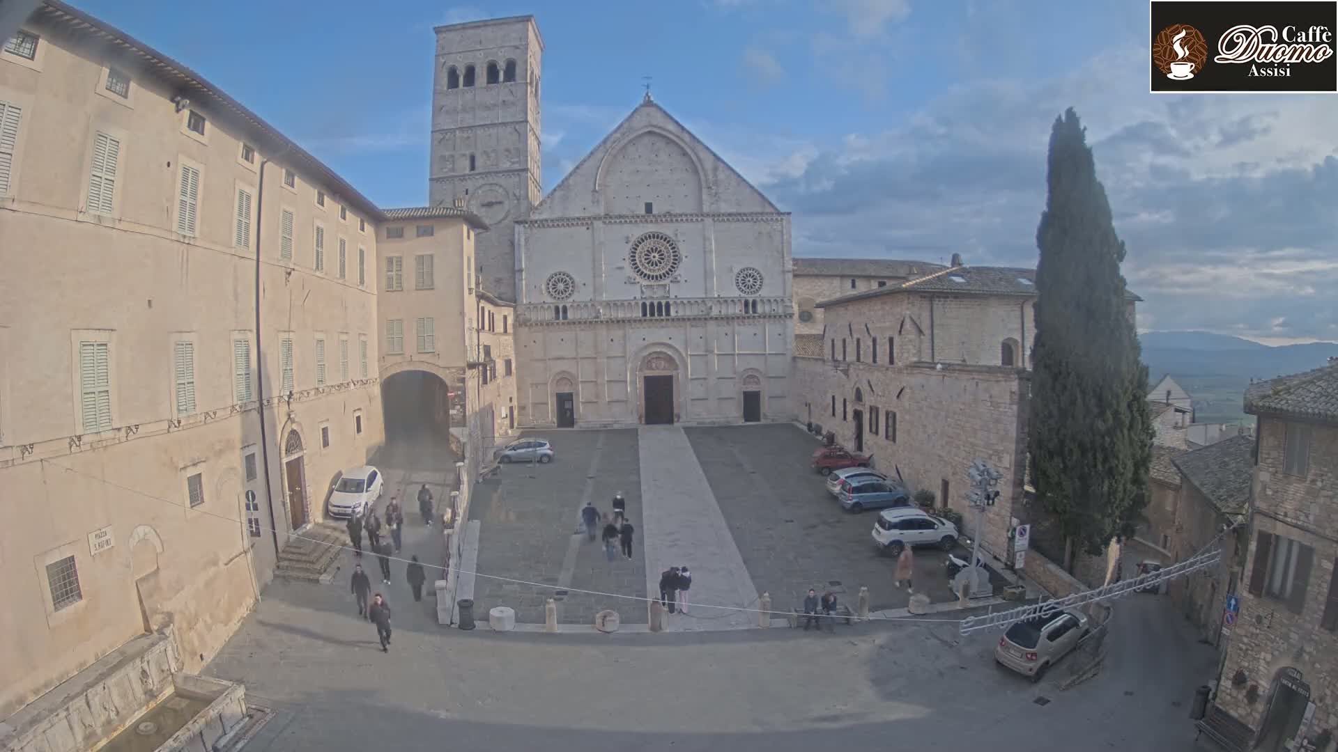 A large stone cathedral stands in a town square under a partly cloudy sky, with several people and cars scattered around the plaza.