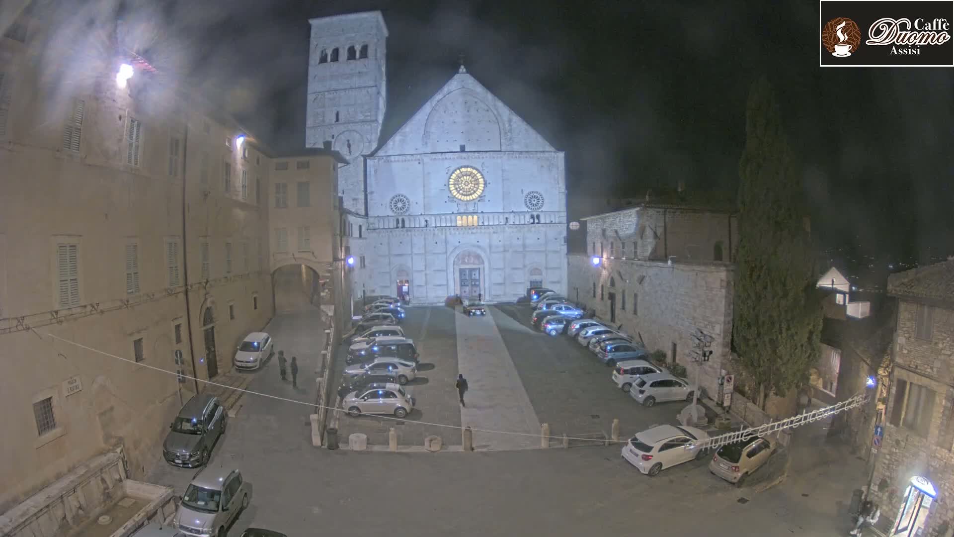 At night, a large cathedral facade is illuminated, with a square in front containing parked cars and a single pedestrian.