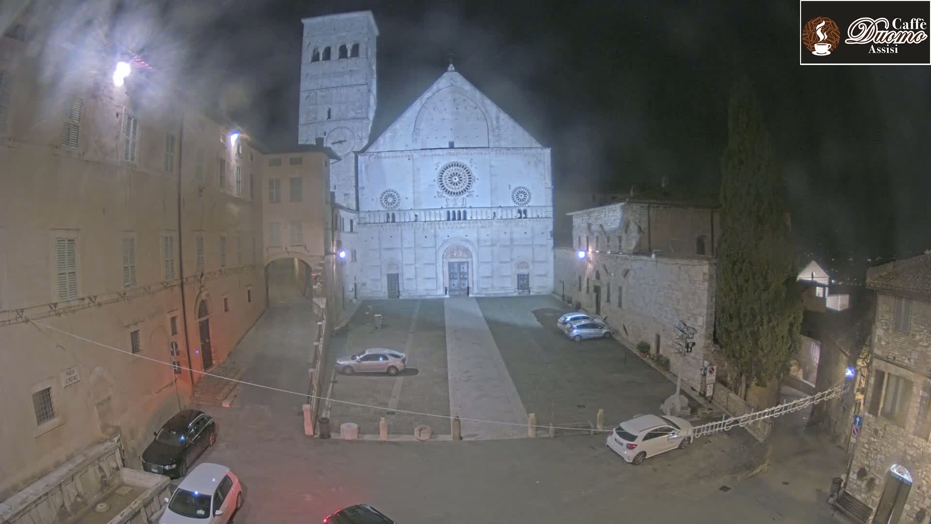 A nighttime view of a large cathedral in a town square, with several cars parked and a hazy atmosphere.