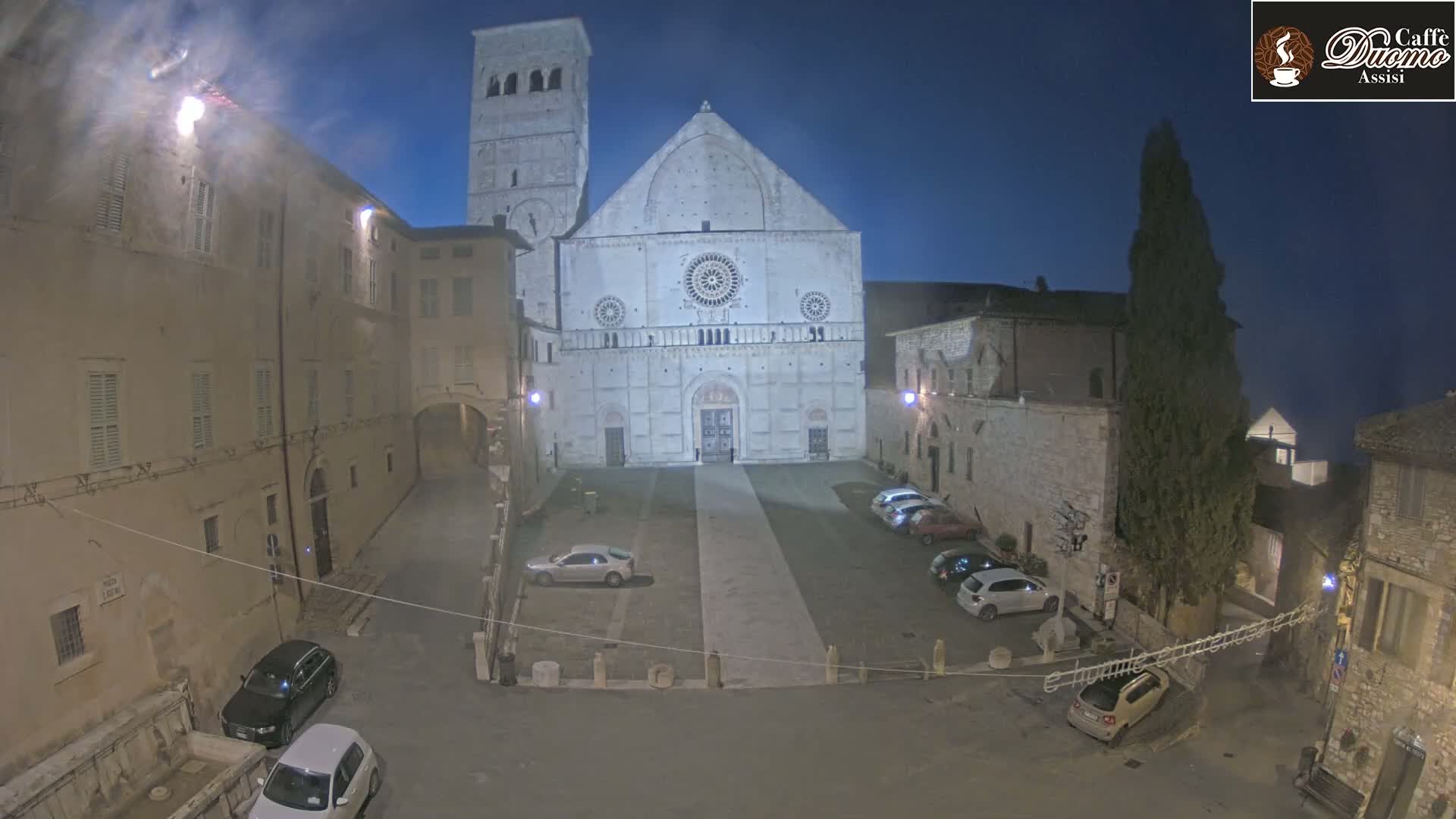 A large, light-colored cathedral stands in a dimly lit square at night, surrounded by stone buildings and a few parked cars.