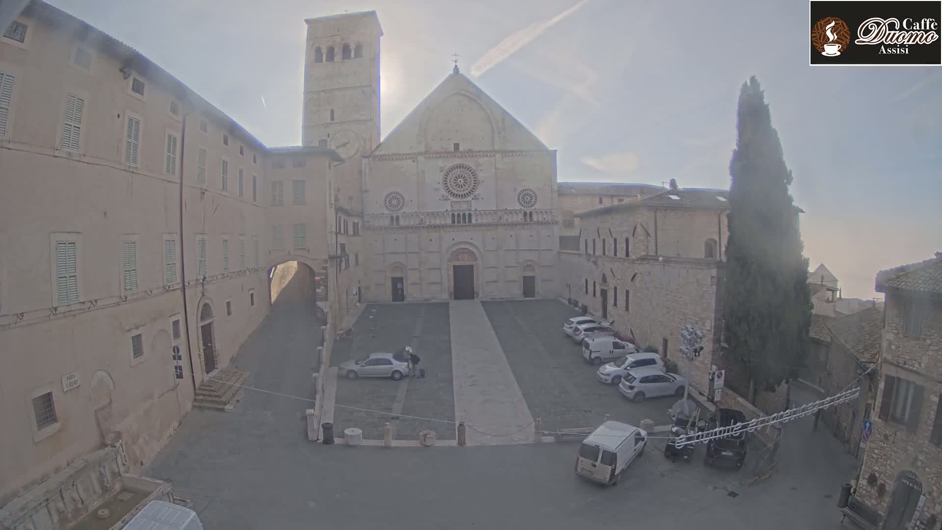 A large stone cathedral stands in a plaza with several parked cars under a hazy, sunny sky.