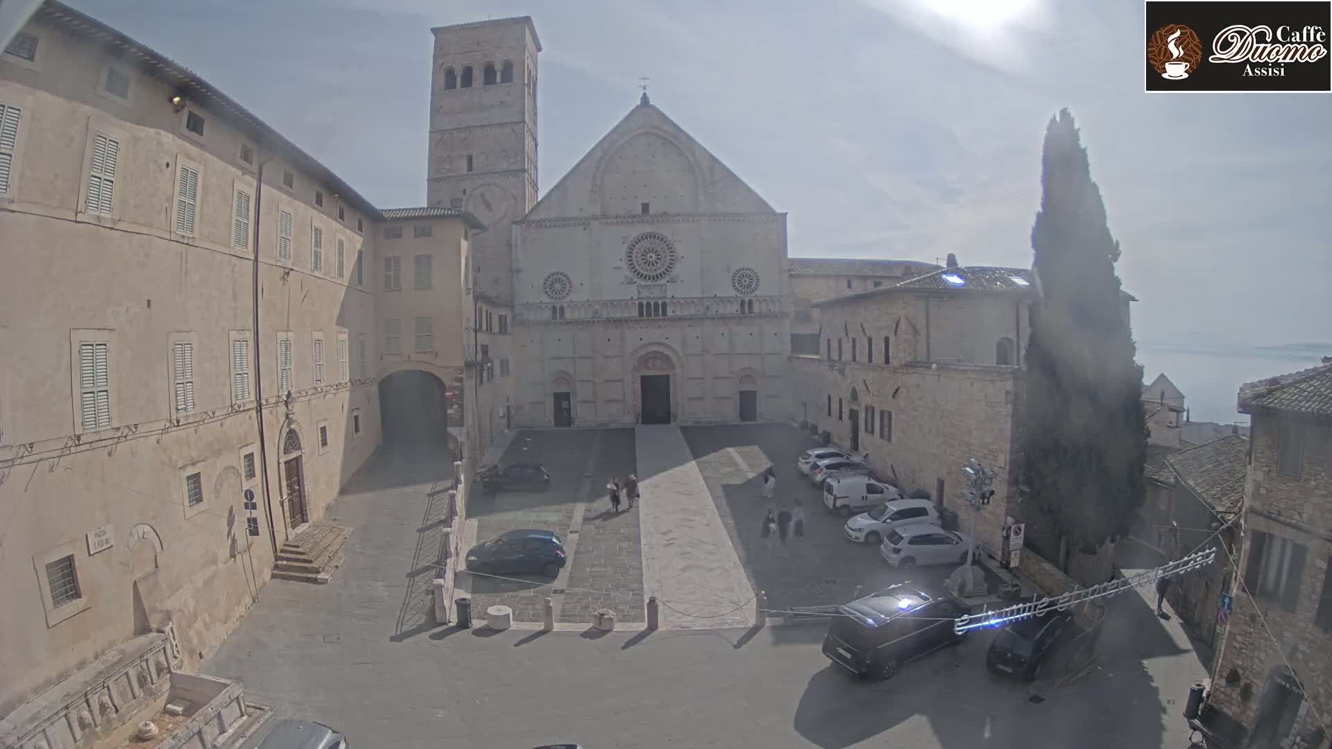 A large stone cathedral stands in a town square on a mostly sunny day, with several cars parked nearby and a few people walking.
