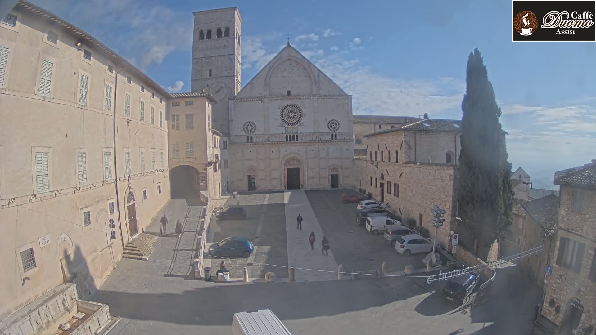 A sunlit square with several parked cars is centered around a large stone cathedral, flanked by adjacent buildings under a mostly clear sky.