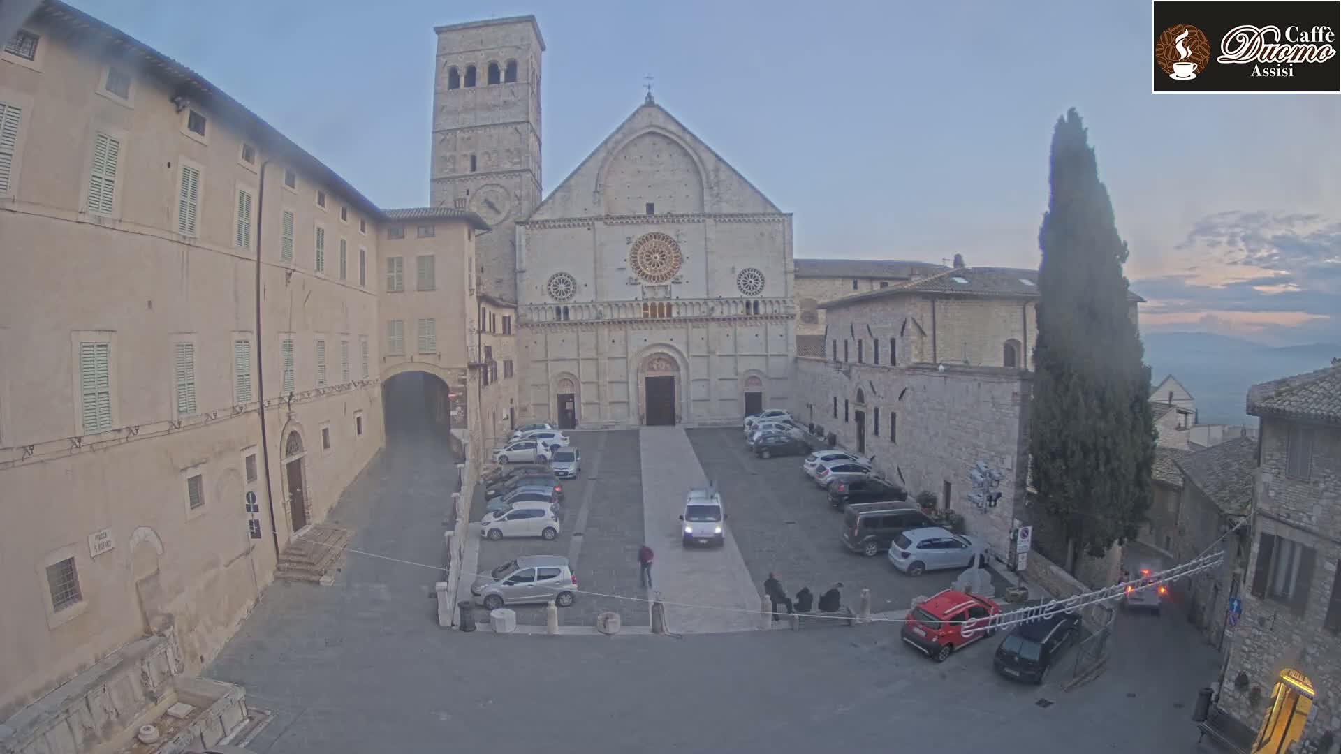 A large stone cathedral sits in a town square with several parked cars under a dusky sky.