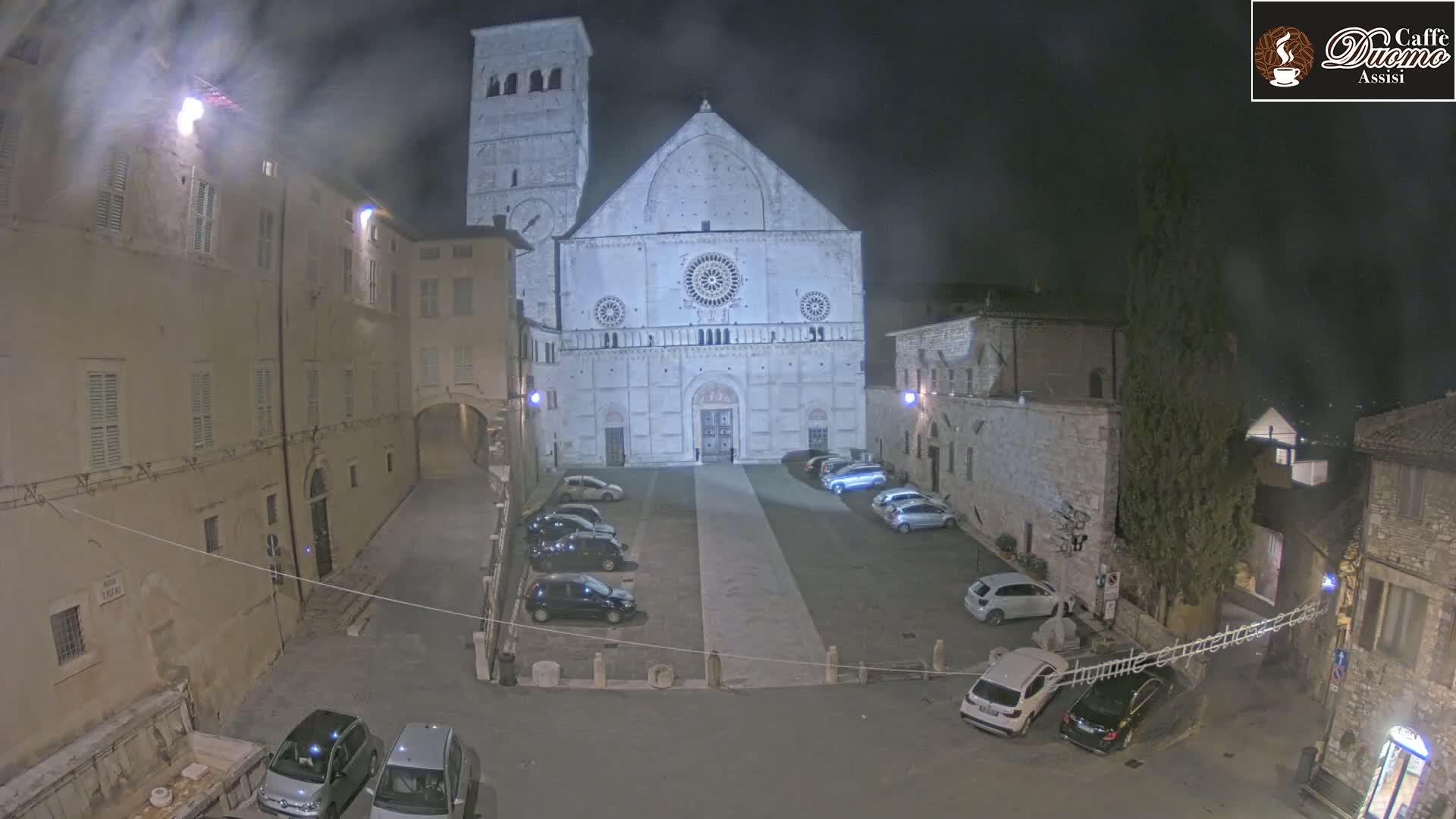 A nighttime view of a large, light-colored cathedral facade in a city square, with several cars parked in the square and surrounding buildings.