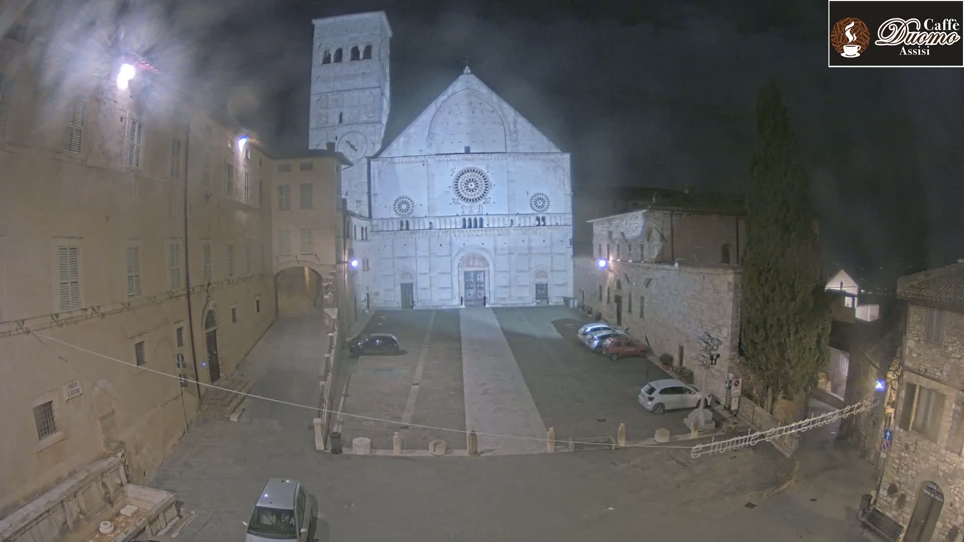 A nighttime view of a cathedral in a town square, illuminated by streetlights, with a few cars parked.