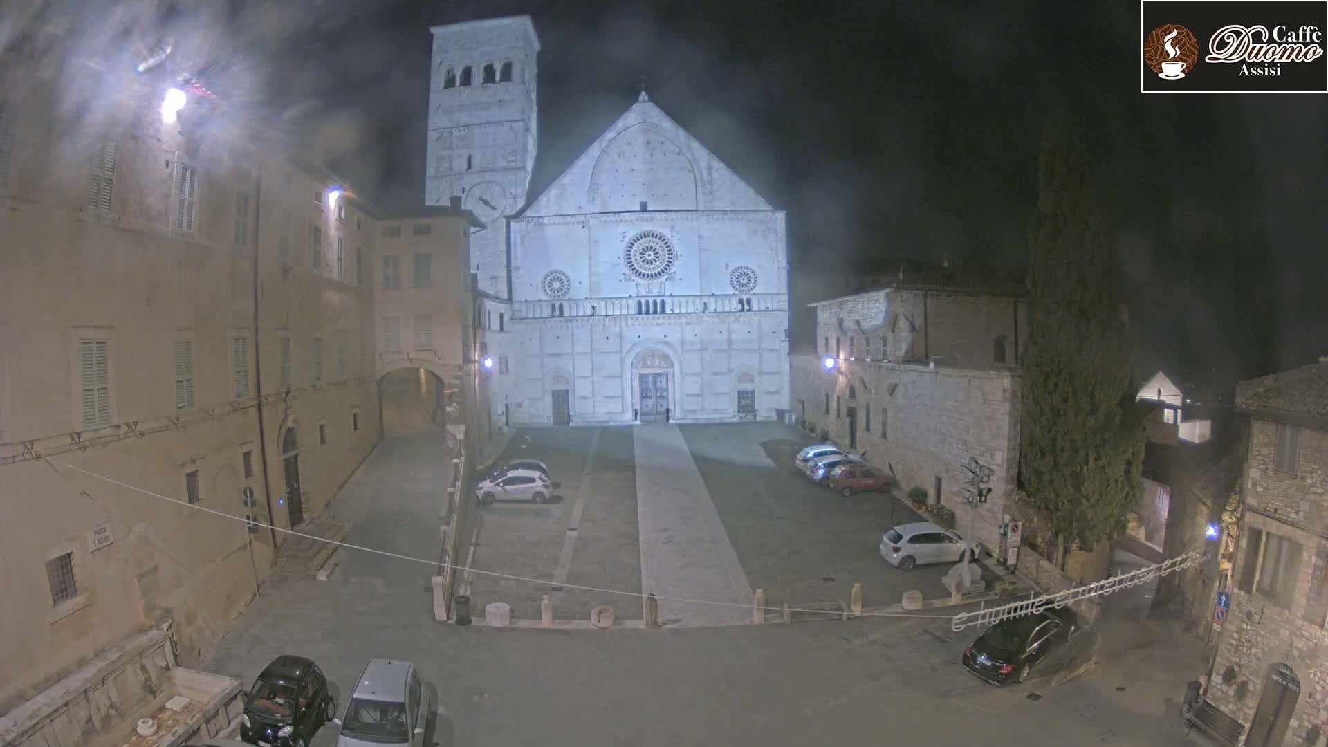 A nighttime view of a large stone cathedral in a town square with several parked cars under hazy conditions.