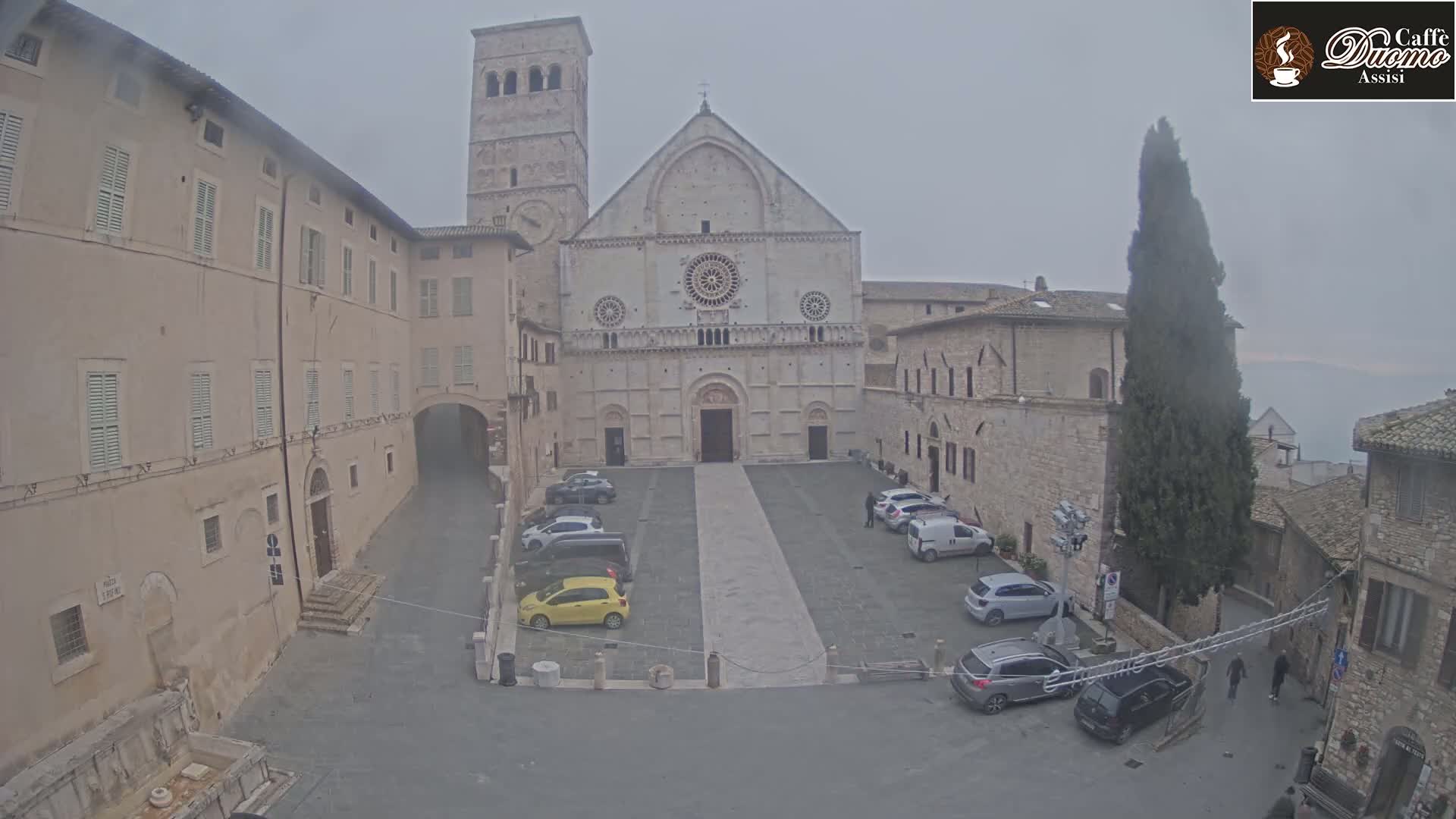 A stone cathedral and surrounding buildings are visible in a square with several parked cars on an overcast day.