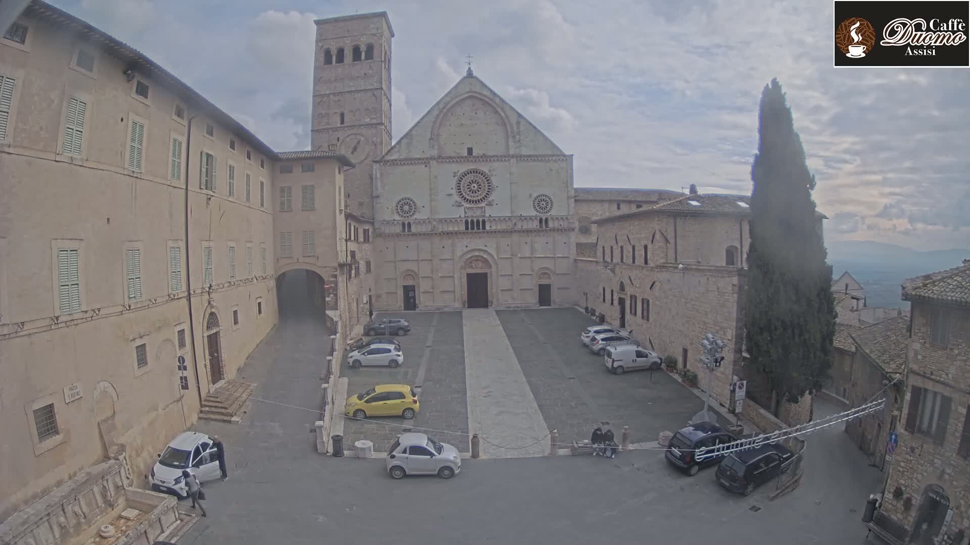A large stone cathedral stands in a town square with several cars parked, under an overcast sky.