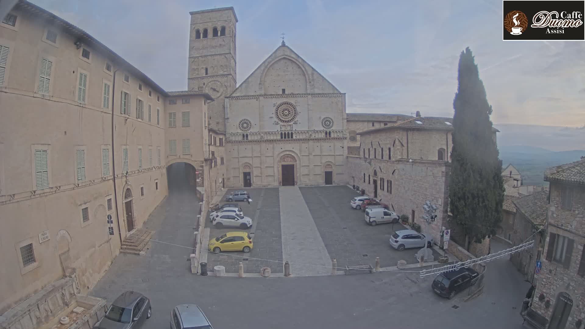 A large stone cathedral with a tall bell tower sits in a town square with several parked cars under an overcast sky.