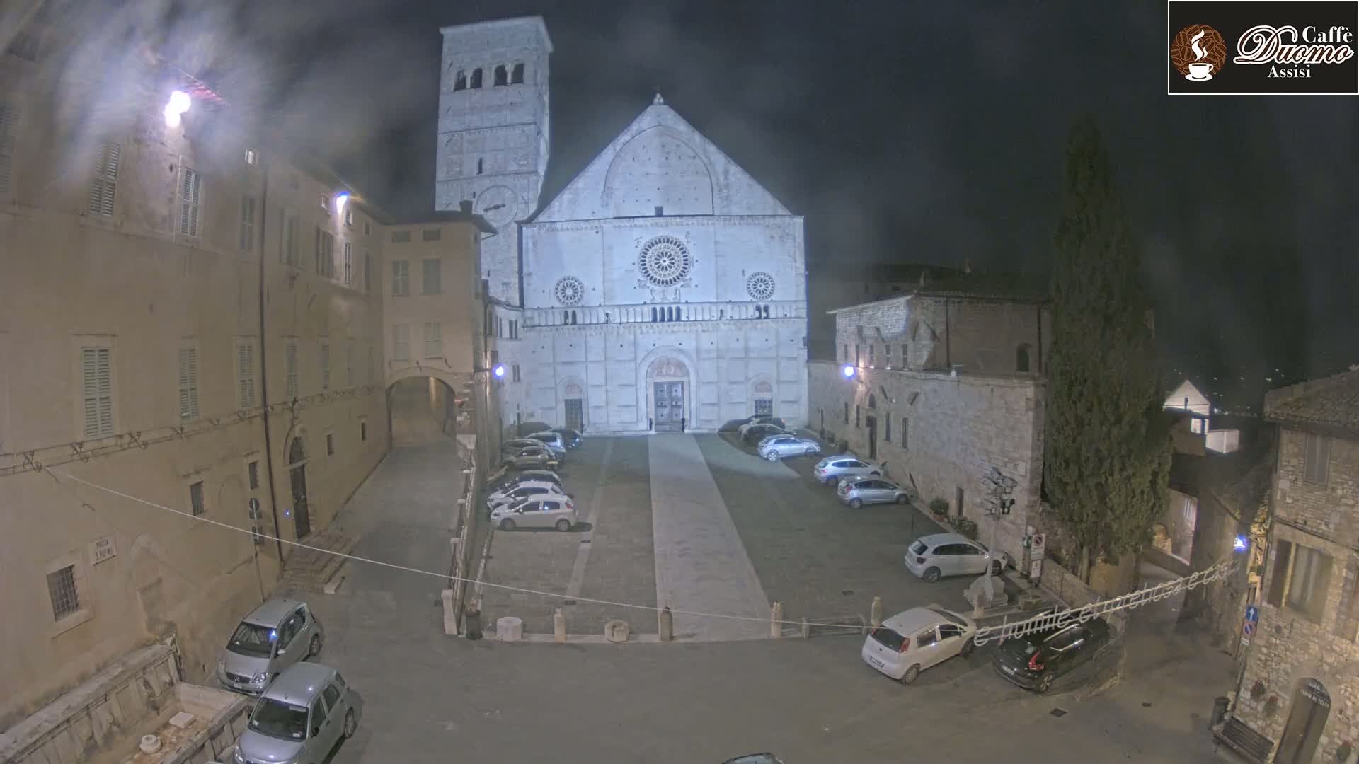 A large stone cathedral is prominently featured in a nighttime, city square with several parked cars under a dark, possibly cloudy sky.