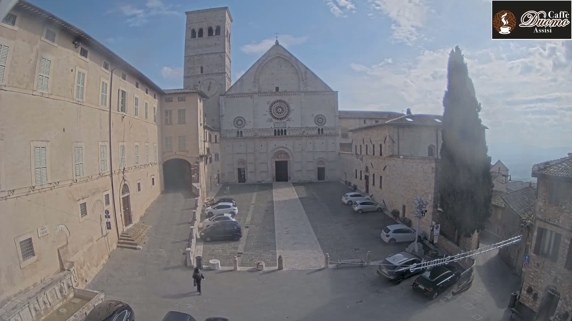 A large stone cathedral sits in a town square with several cars parked around it, under a partly cloudy sky.