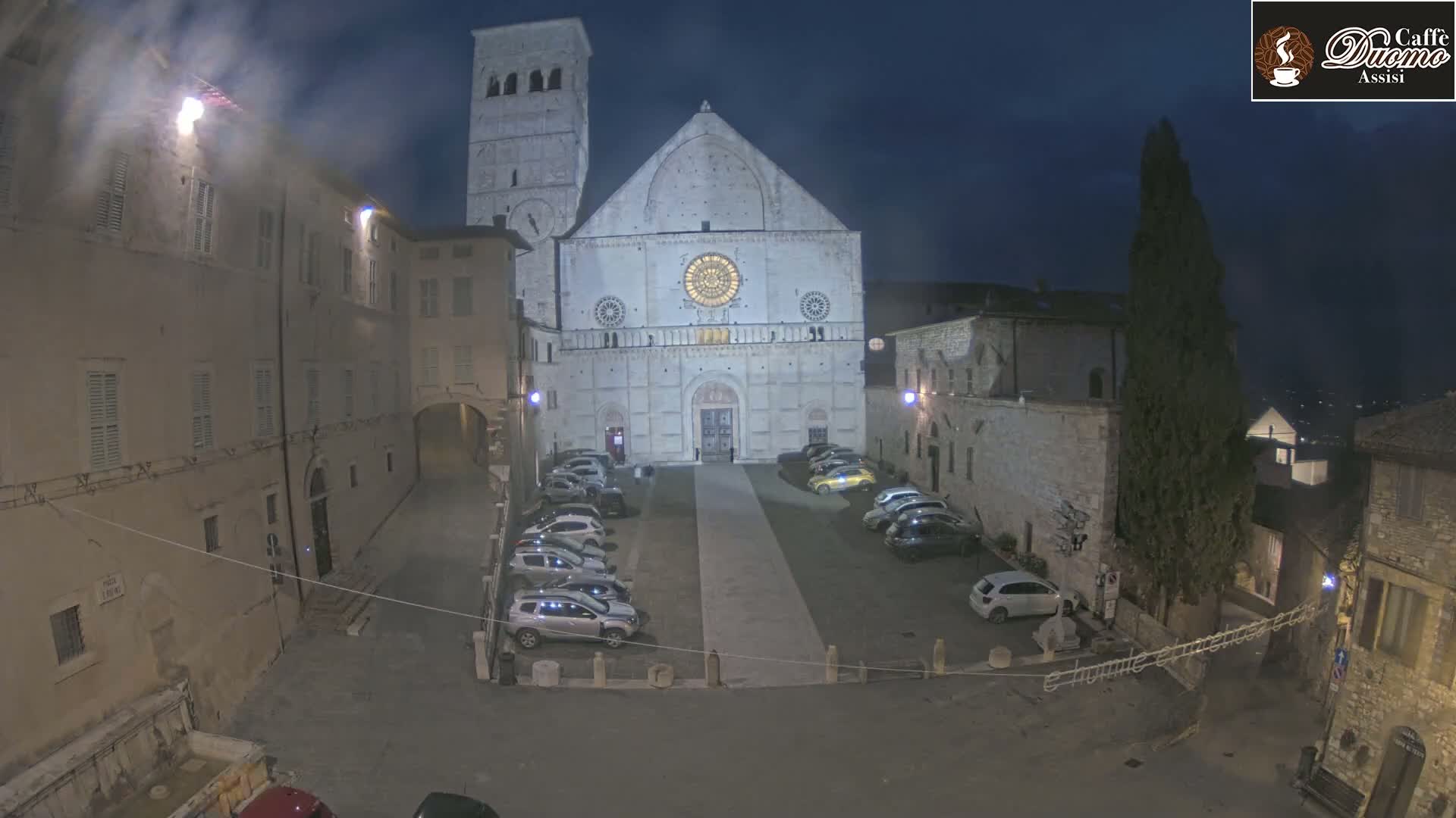 A large stone cathedral is illuminated at night in a mostly empty town square with cars parked on either side of a walkway.