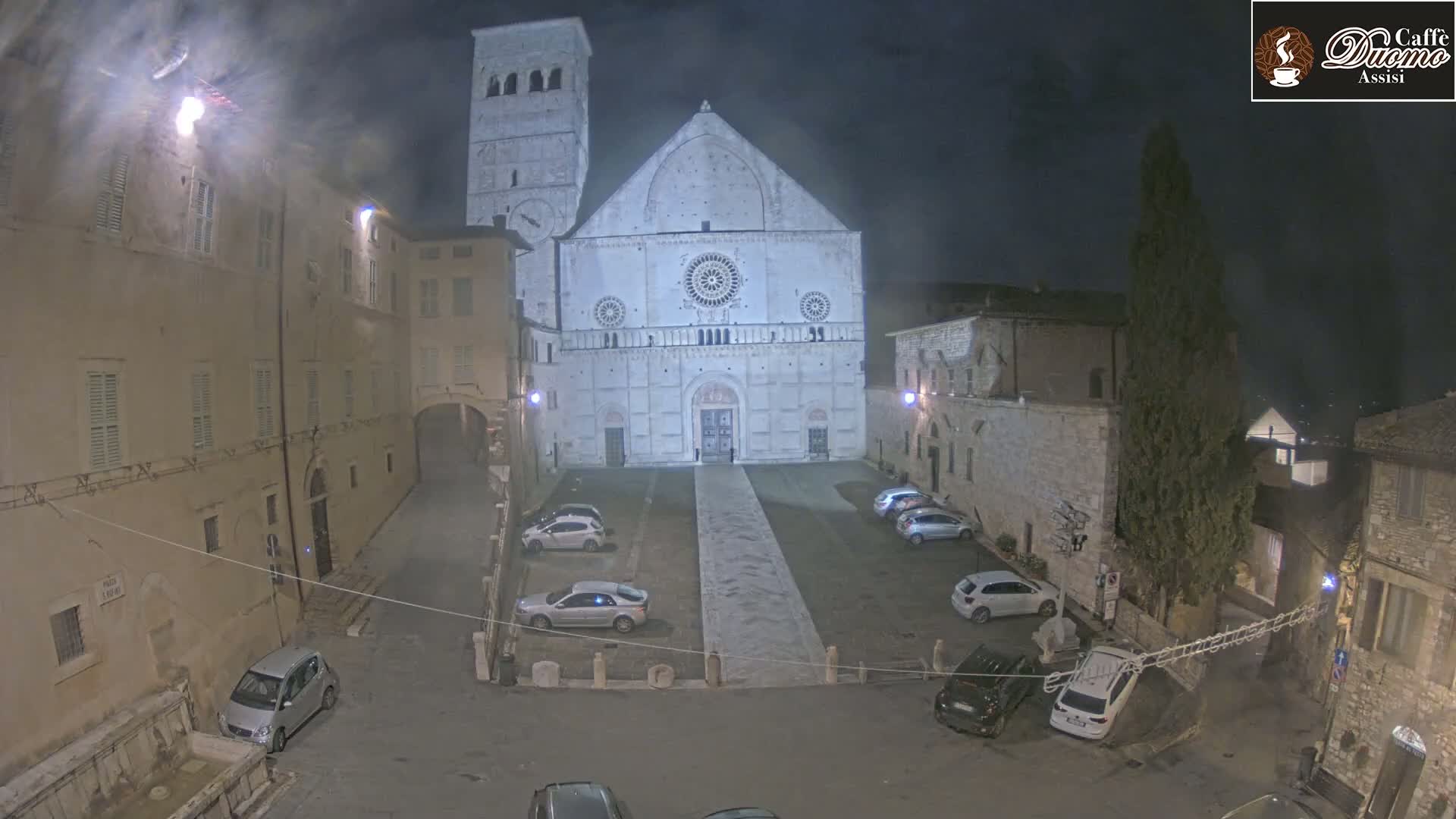 At night, a large stone cathedral faces a mostly empty square with several cars parked, surrounded by old stone buildings under a dark, cloudy sky.