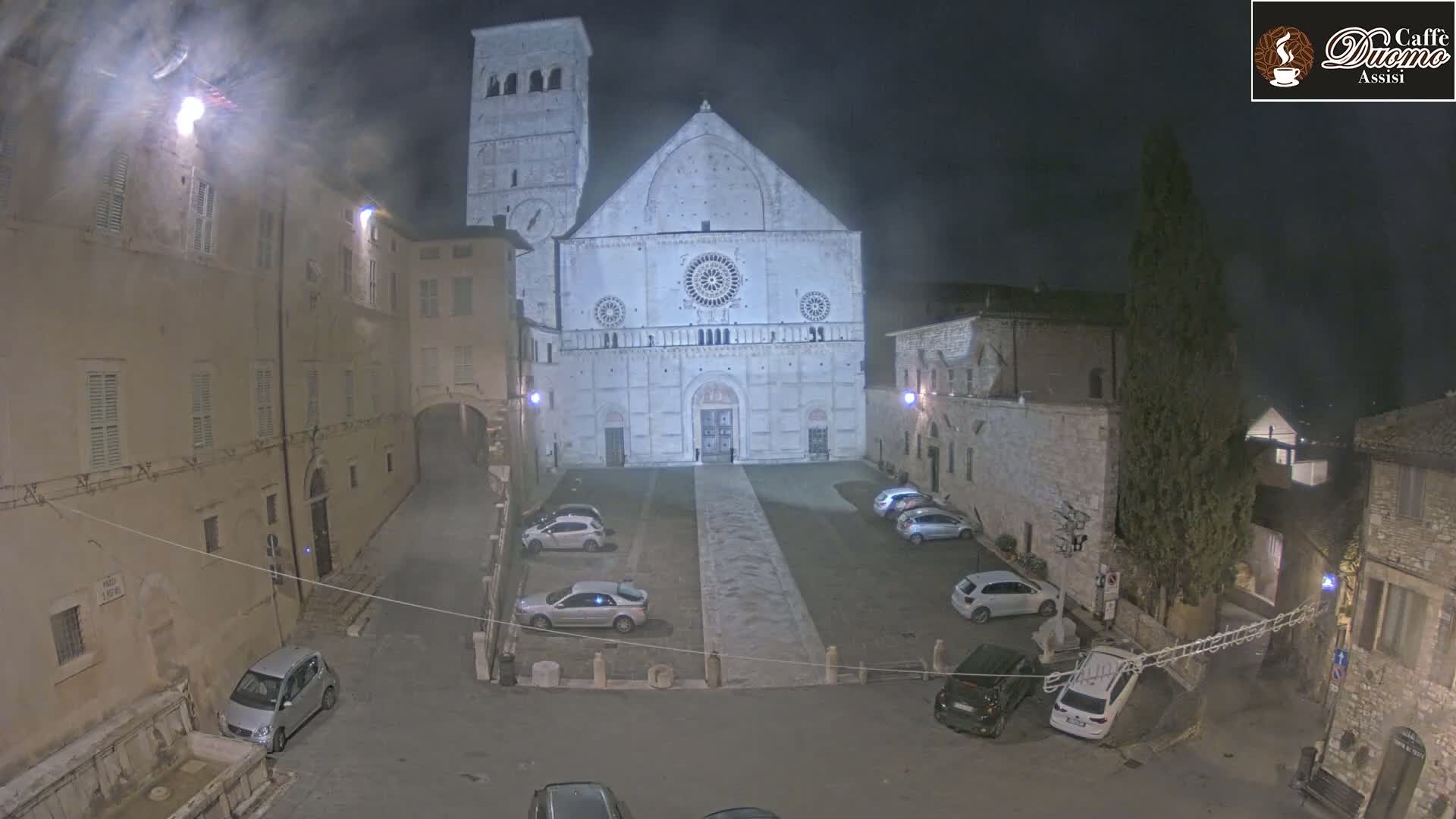 A large, light-colored cathedral stands in a dimly lit, empty square at night, surrounded by older stone buildings and parked cars.