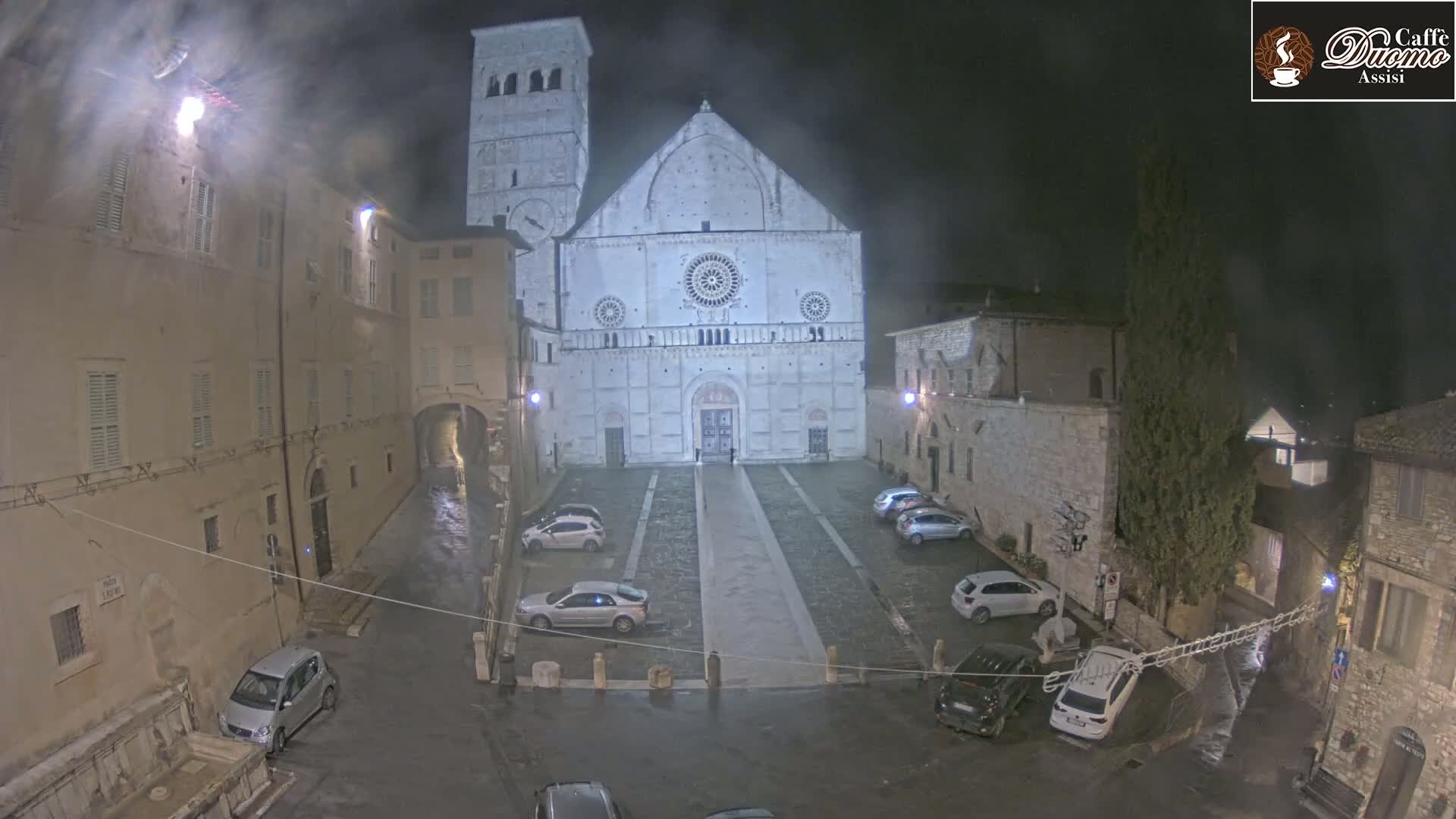 A large, light-colored cathedral stands in a dimly lit, rain-slicked town square at night, surrounded by parked cars and older buildings.