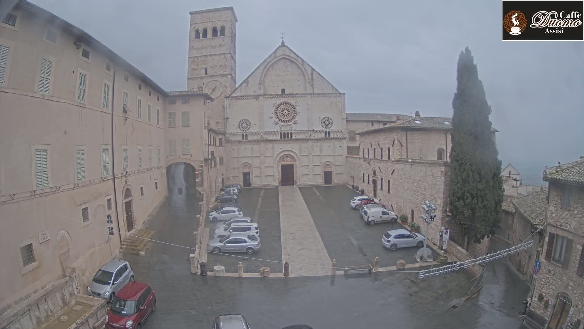 A large stone cathedral stands in a town square with cars parked on wet pavement under an overcast sky.