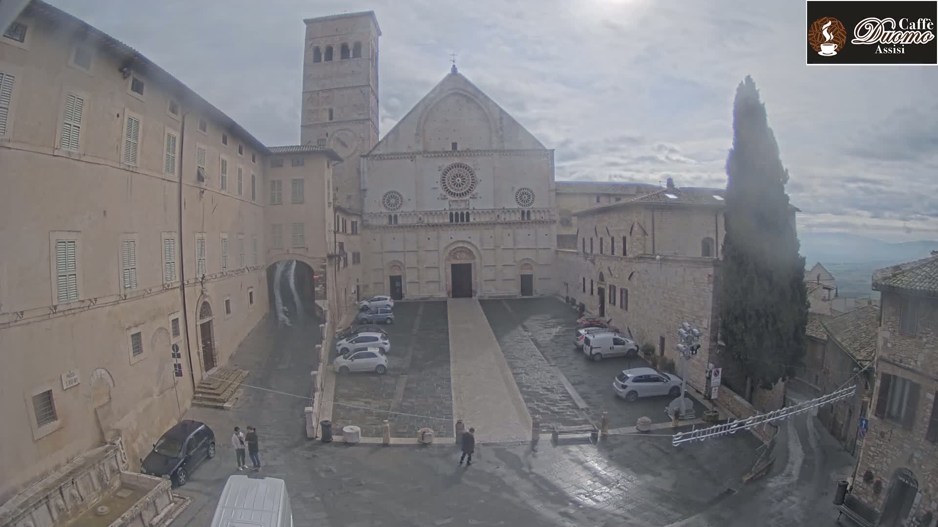 A large stone cathedral sits in a town square, surrounded by other stone buildings under an overcast sky.