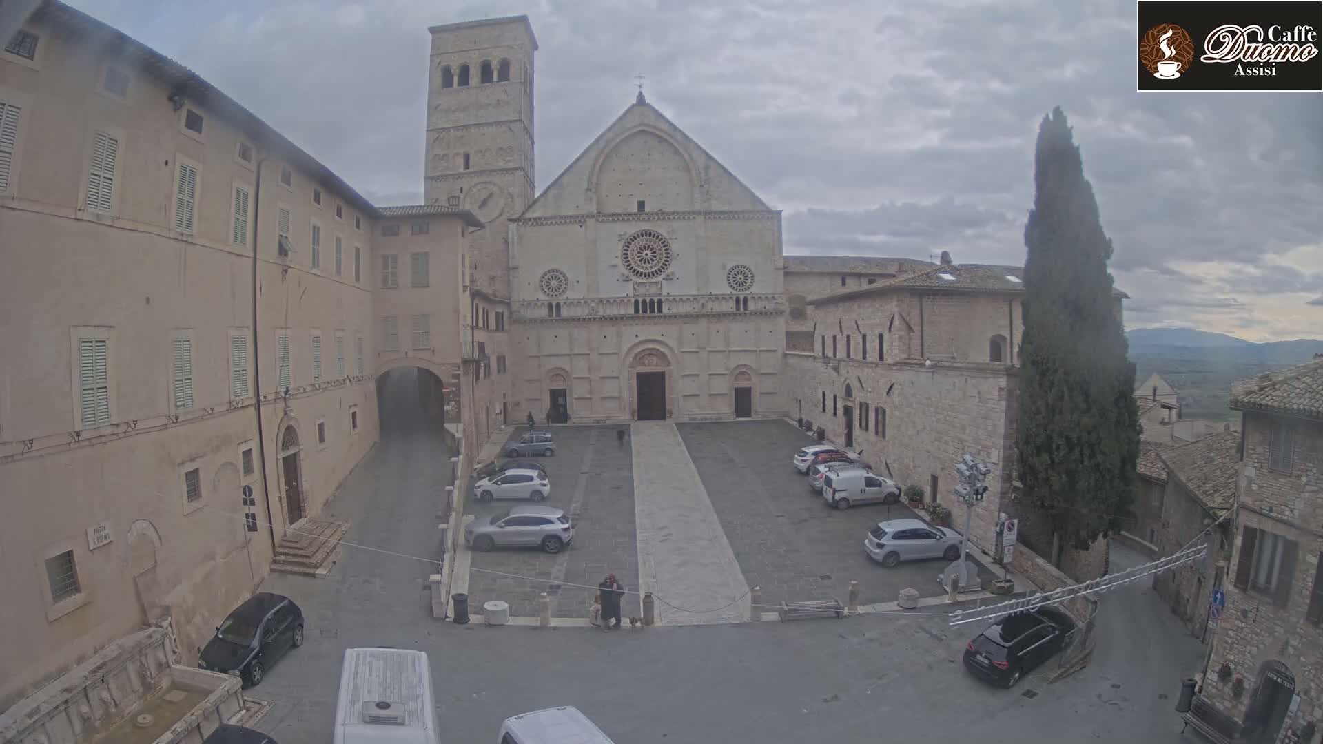 A large stone cathedral sits in a town square with several cars parked, under an overcast sky.