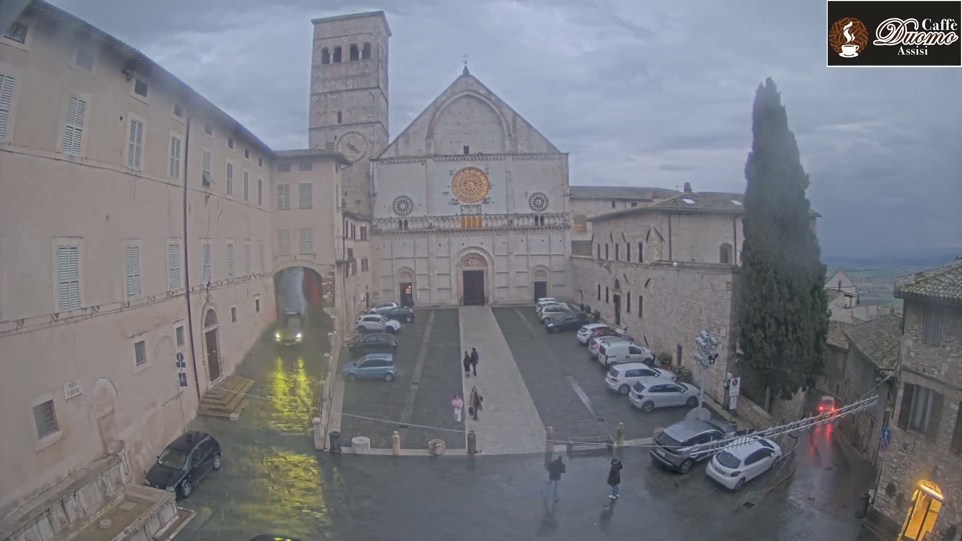 A large stone cathedral stands in a town square, surrounded by buildings and parked cars under an overcast sky, with rain falling.