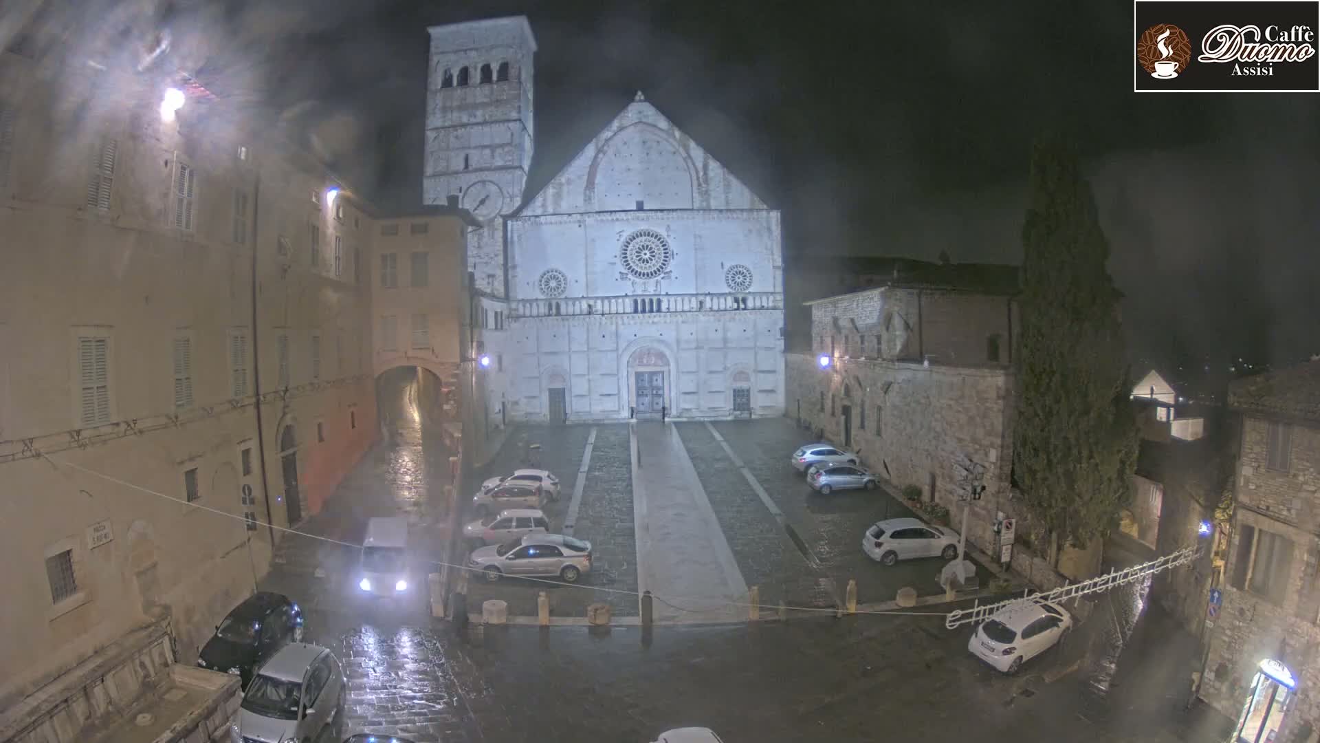 A large stone cathedral is situated in a town square at night during a light rain, surrounded by parked cars and buildings.
