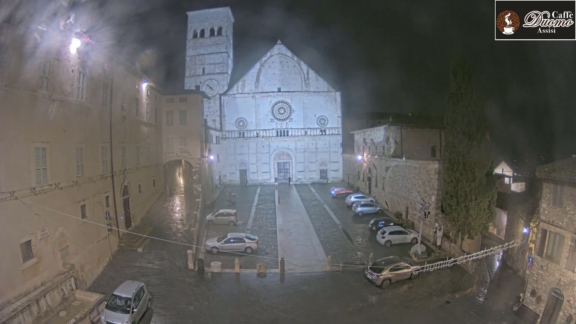 A large stone cathedral stands in a town square at night during a light rain, surrounded by old buildings and a few parked cars.