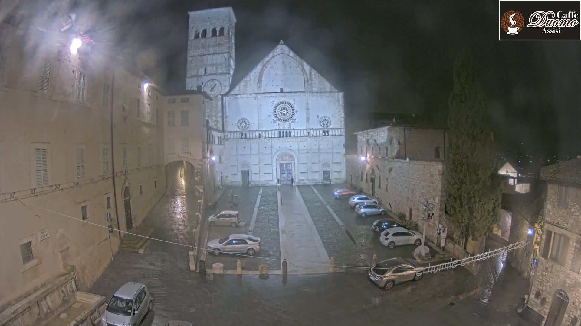 A large stone church stands in a town square at night during a light rain, with several cars parked in the square and wet streets surrounding the church.