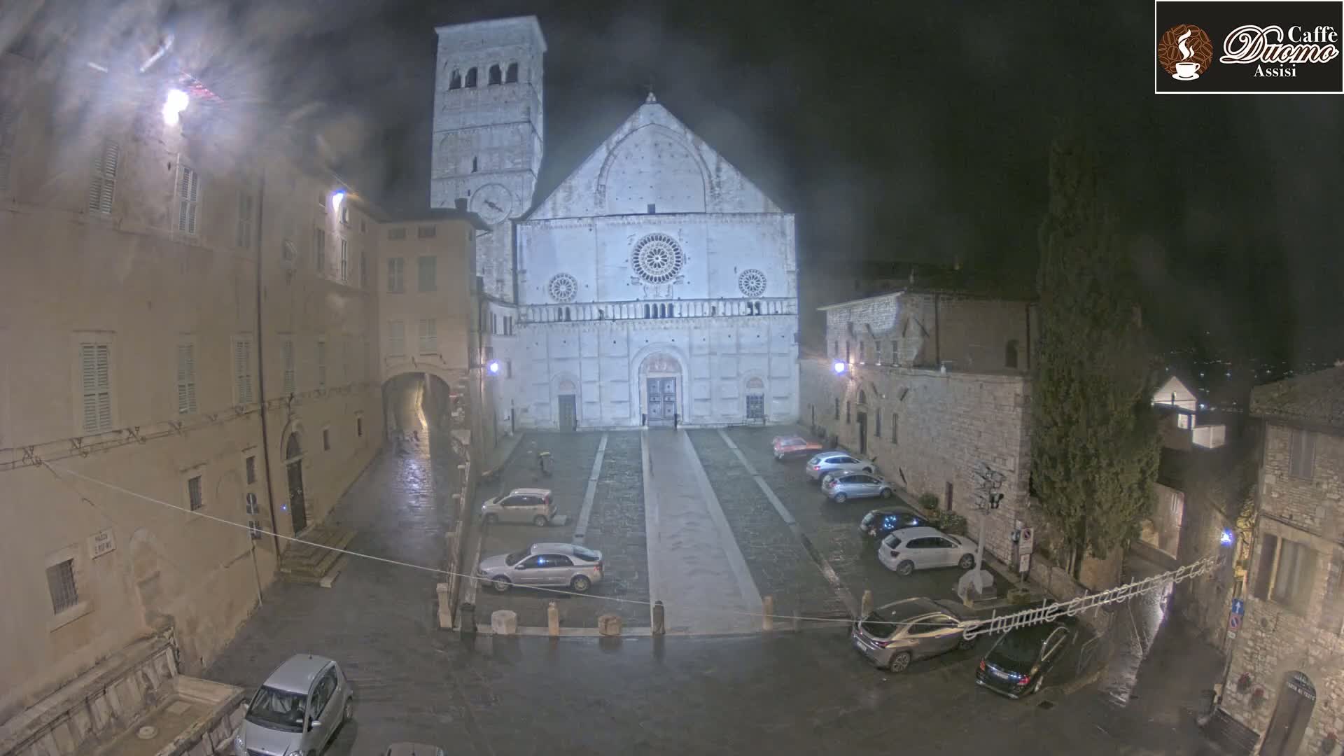 A nighttime, rain-soaked view of a large cathedral in a town square, with cars parked around the perimeter and buildings lining the square.