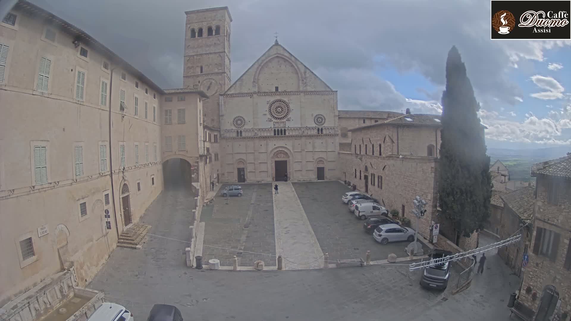 A large stone cathedral faces a town square with several parked cars under an overcast sky.