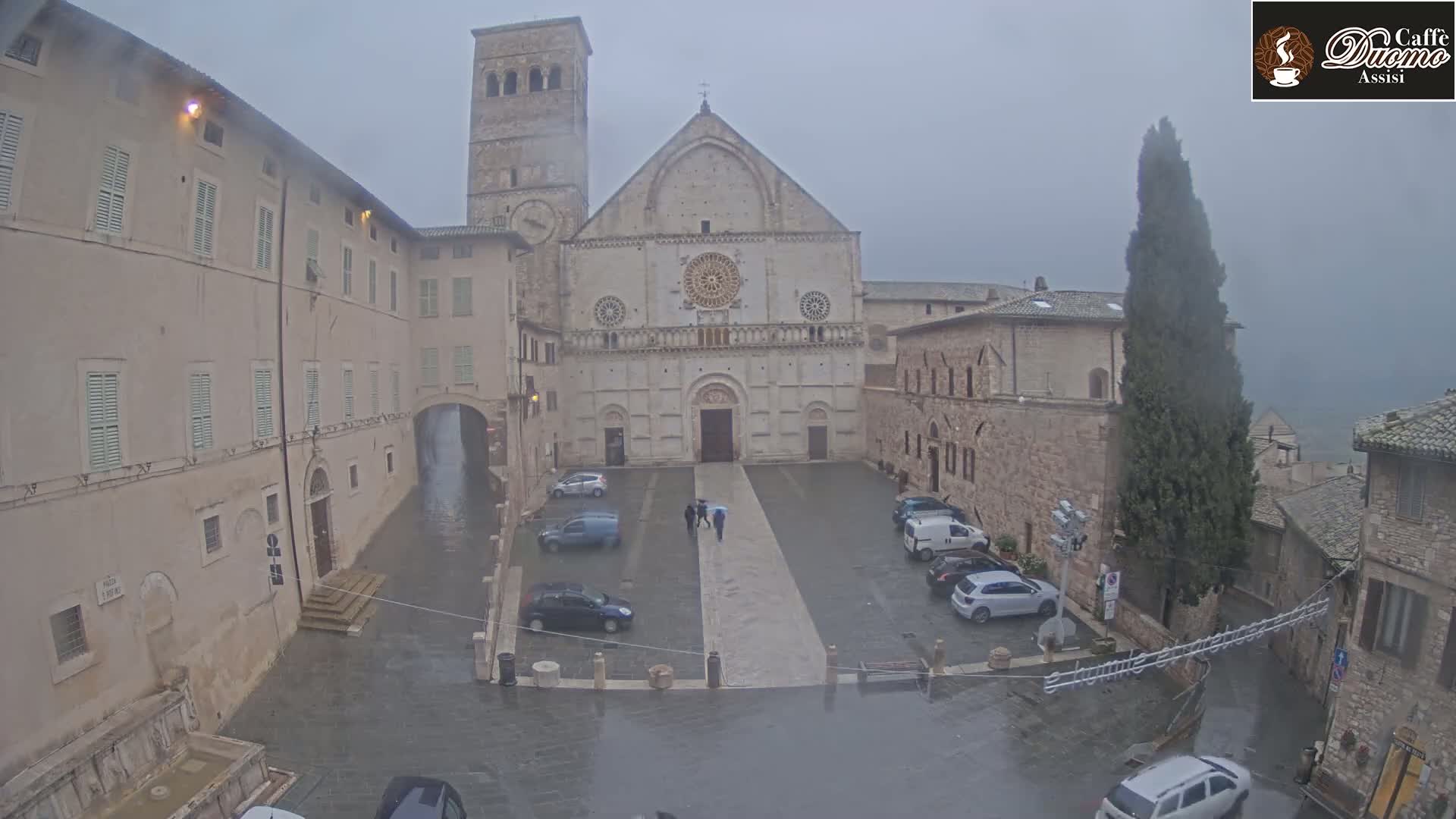 A large stone cathedral stands in a rain-soaked town square, surrounded by old buildings and a few parked cars.